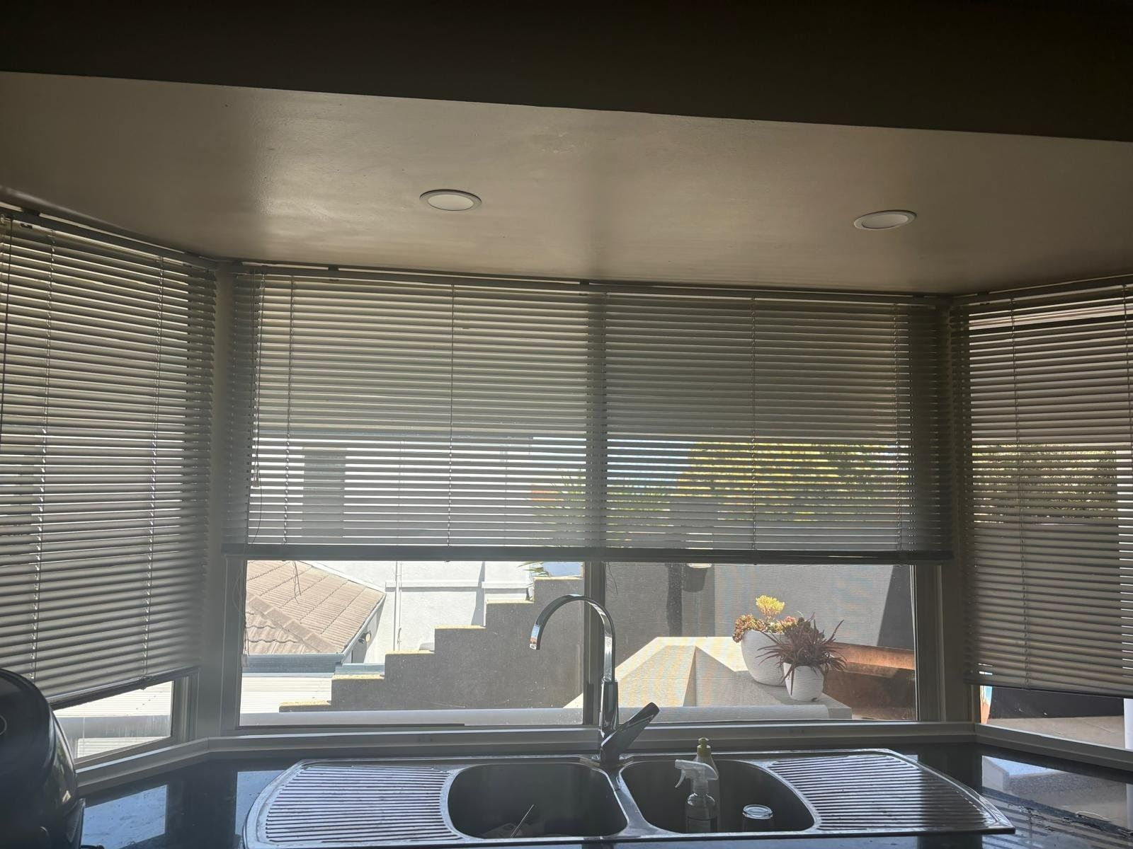 Plantation shutters installed in a kitchen sink beneath a curved window with blinds, looking out onto a patio in Perth, WA.