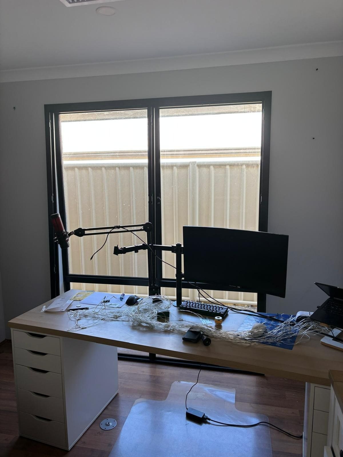 Plantation shutters installed in a messy home office with a desk, monitor, mic arm, and a window with blinds in Perth, WA.