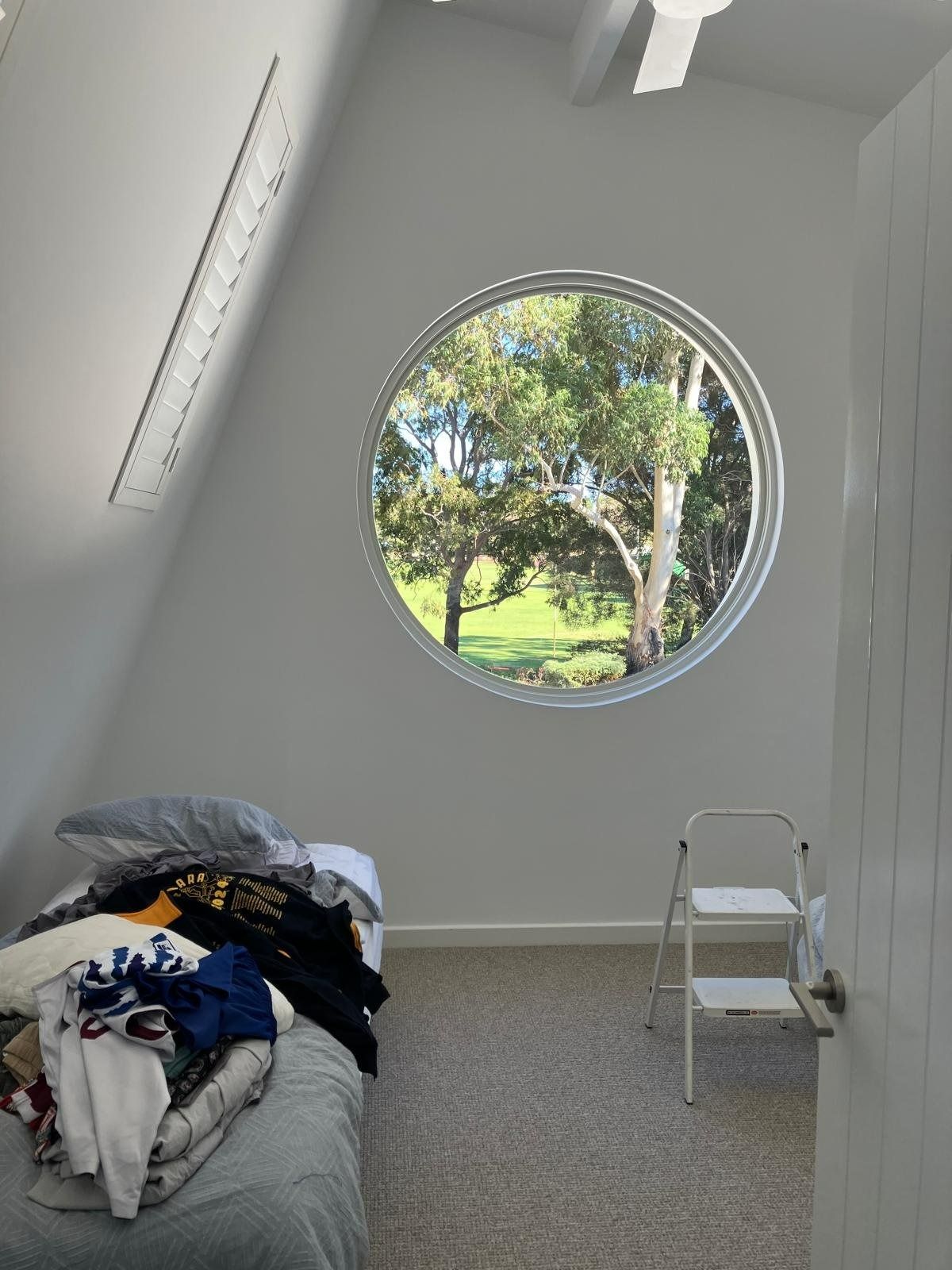 Plantation shutters installed in a minimal bedroom with a round window, slanted ceiling, bed, and small step ladder in Perth, WA.