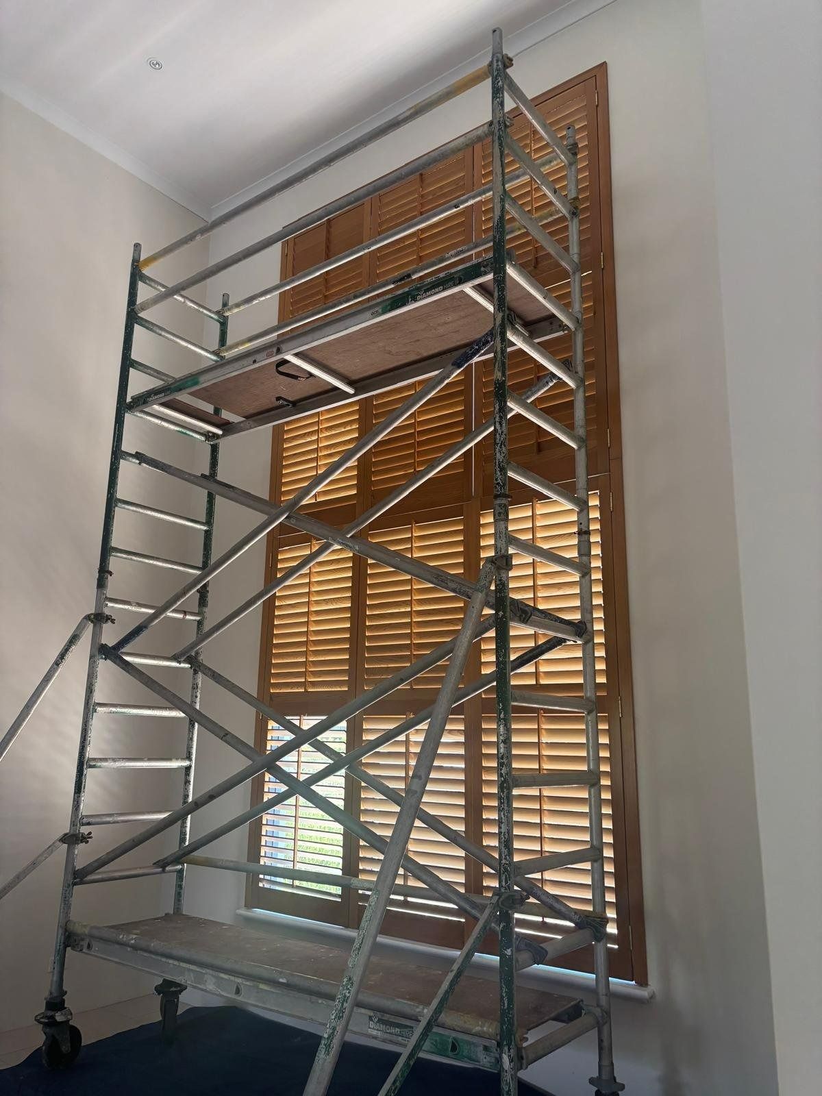 Plantation shutters installed in a scaffolding inside a room around a large window with blinds, against white walls in Joondalup, WA.
