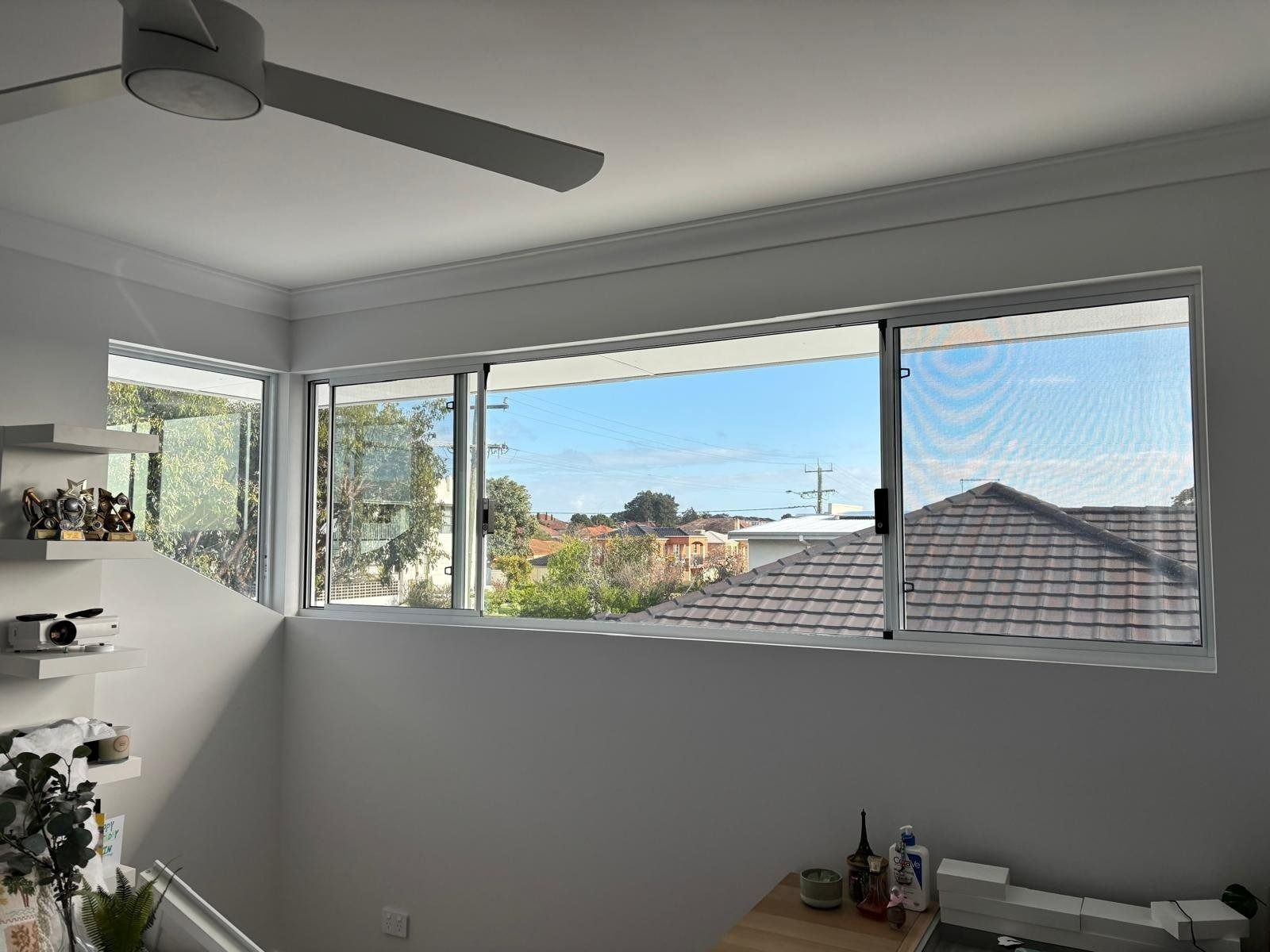 Plantation shutters installed in a bright room with large windows, ceiling fan, and a view of rooftops and blue sky in Perth, WA.