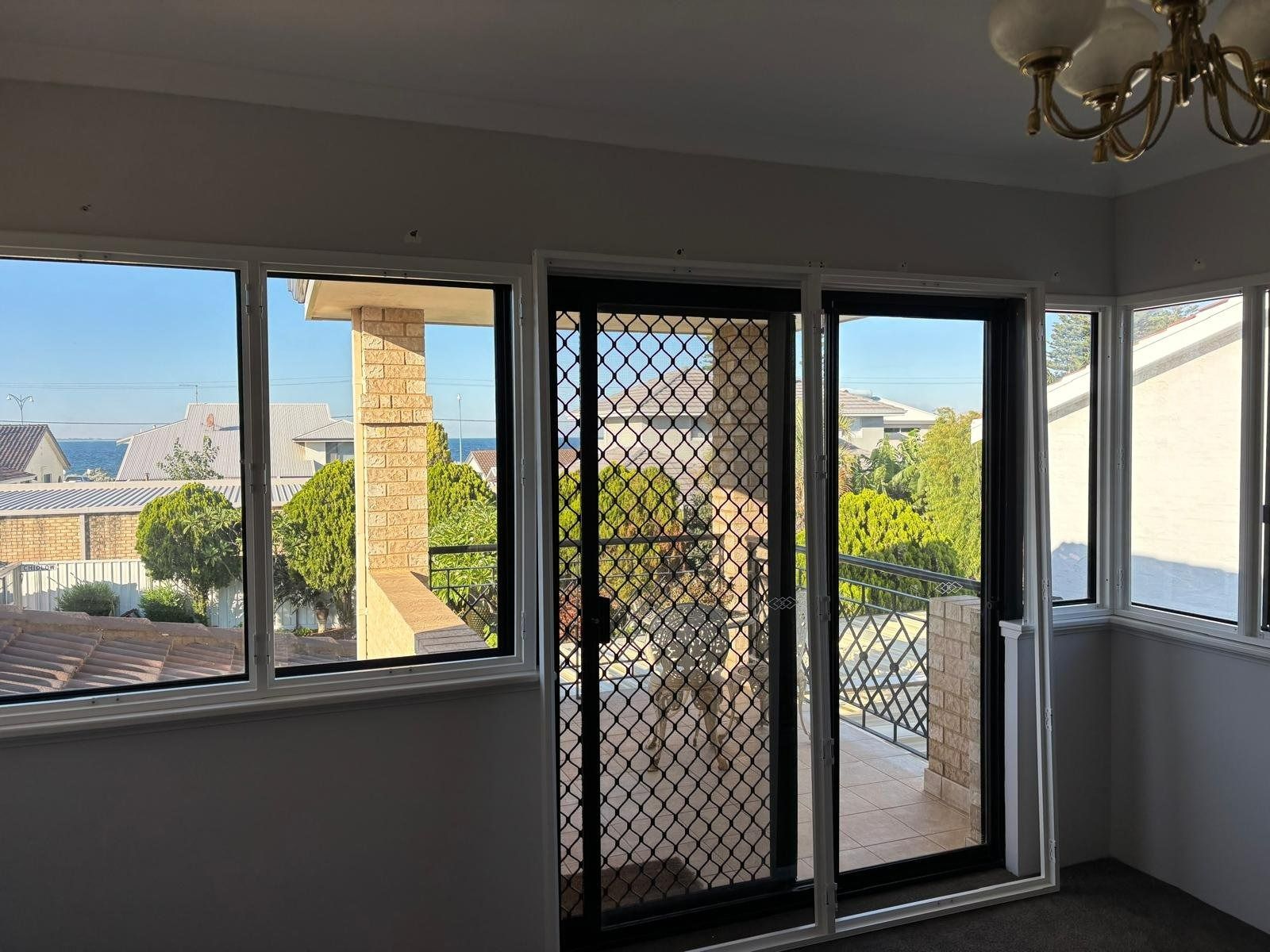 Plantation shutters installed in a bright room with a sliding screen door and windows overlooking a balcony and neighborhood rooftops in Joondalup, WA.