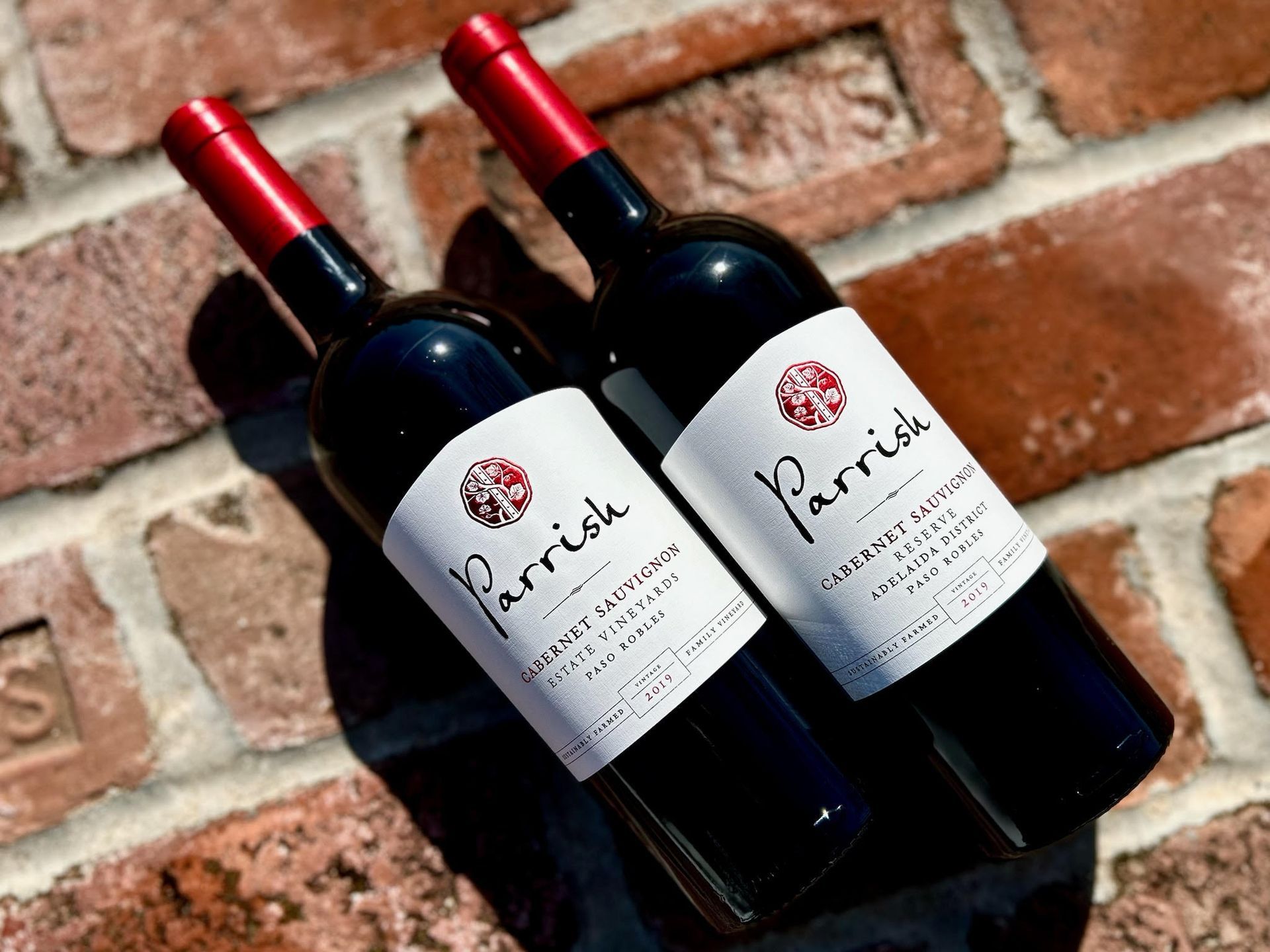 Two bottles of Parish Cabernet Sauvignon wine with red caps and white labels on brick.