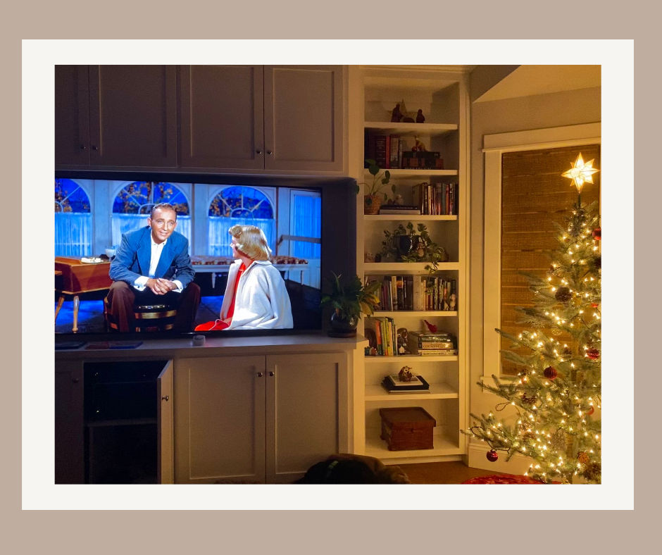 Living room with holiday movie on the television, bookshelf, and a lit Christmas tree at the right.