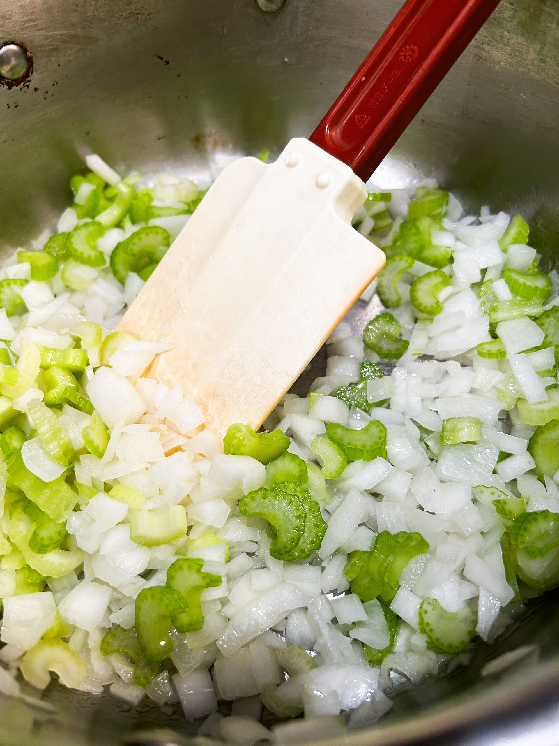 A wooden spatula is stirring onions and celery in a pot.