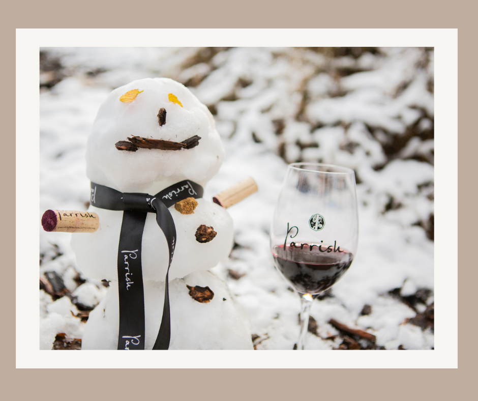 A snowman decorated in Parrish corks and ribbon, alongside a glass of Parrish red wine in a snowy outdoor setting.