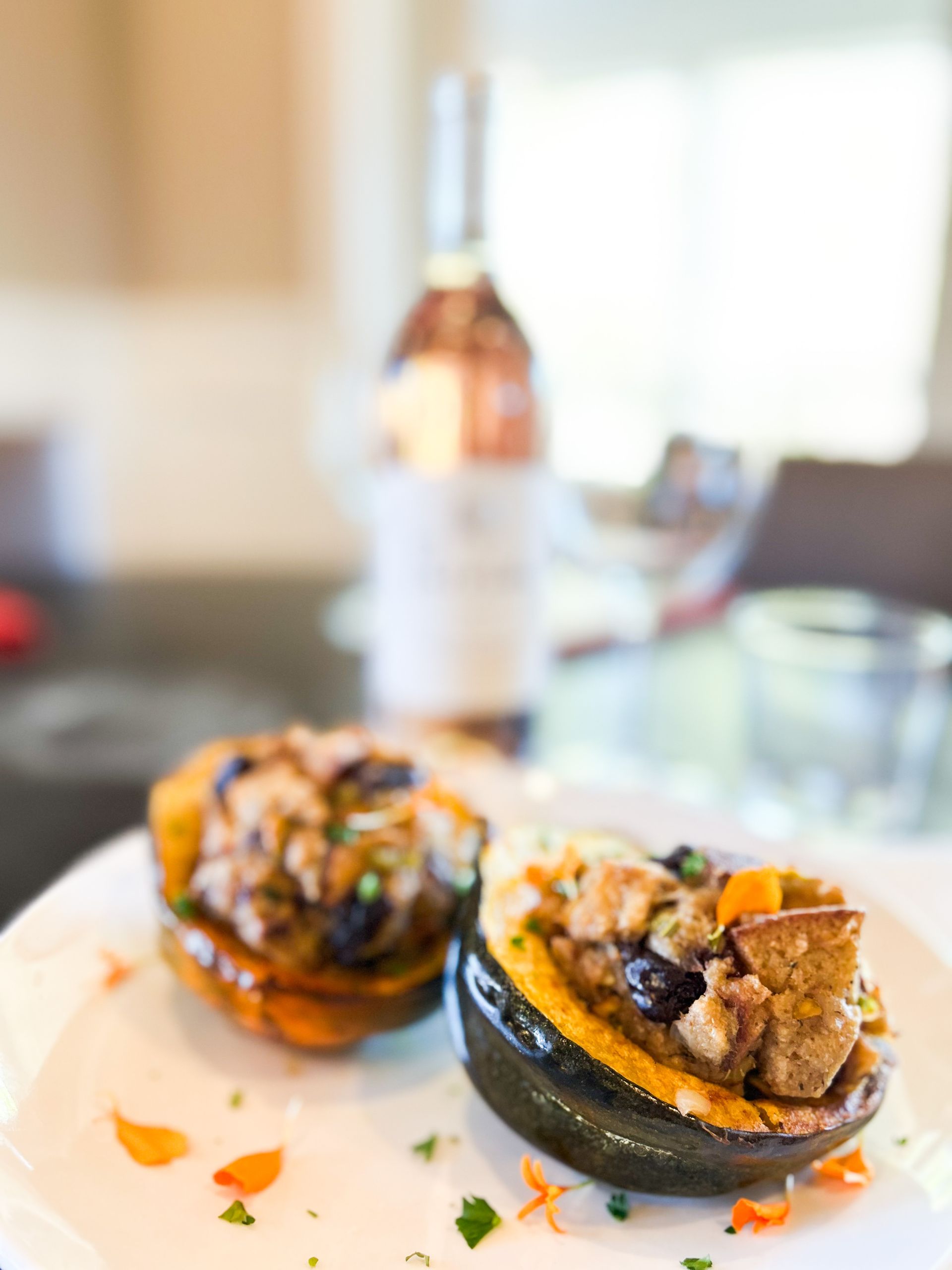 Two stuffed acorn squash on a white plate with a bottle of wine in the background