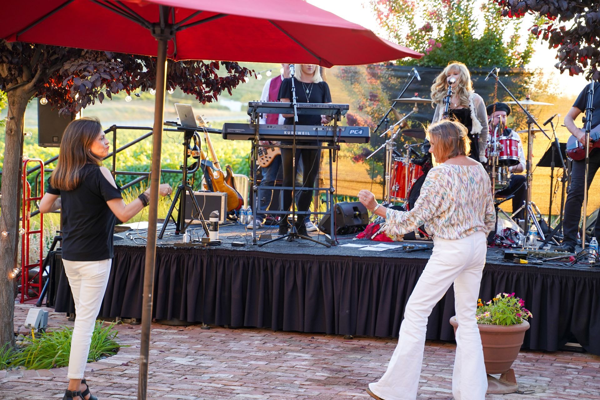 Outdoor band performing on a patio stage under red umbrellas, with two women dancing in the foreground.