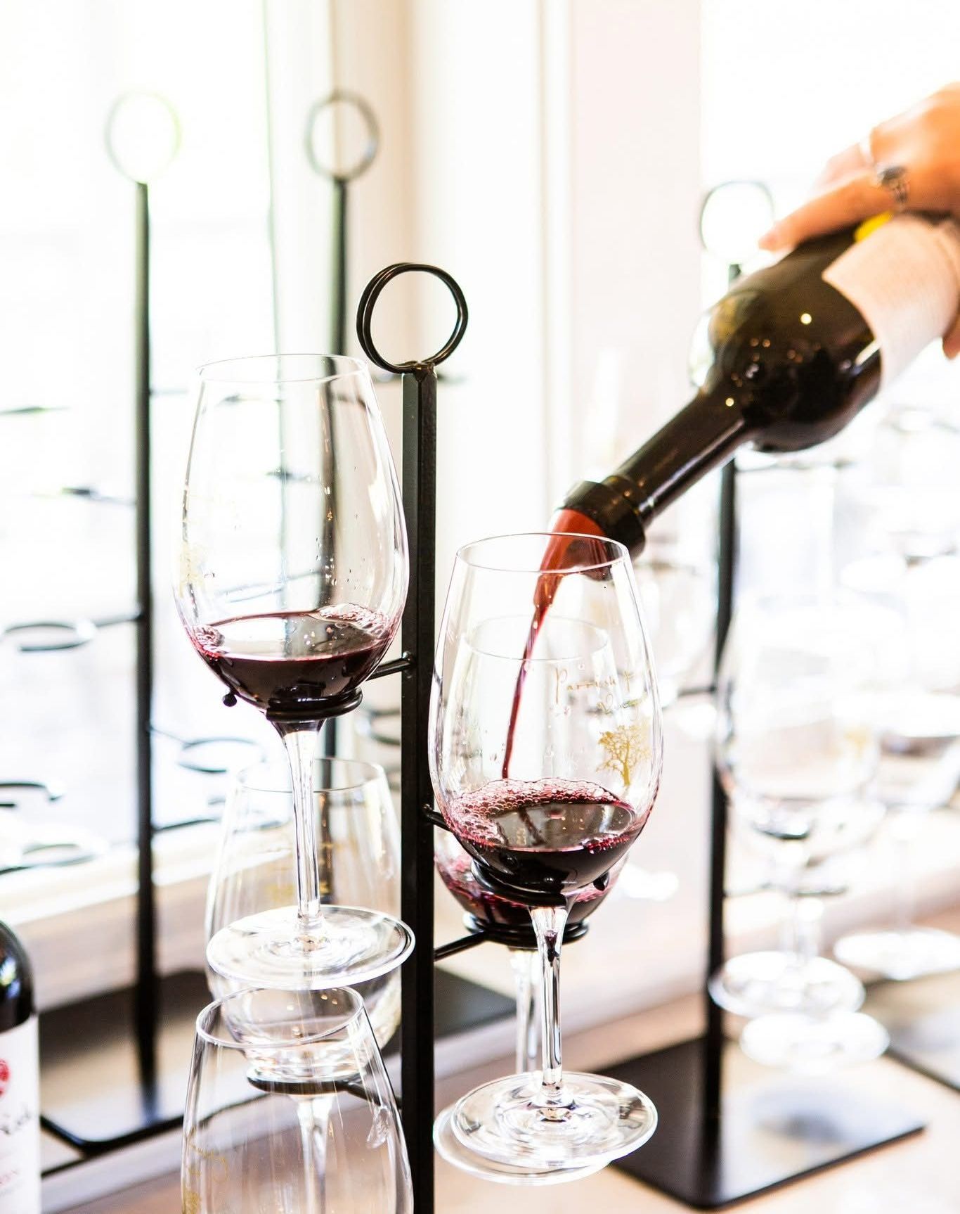 Person pouring red wine into a stem glass in a wine rack with multiple glasses.