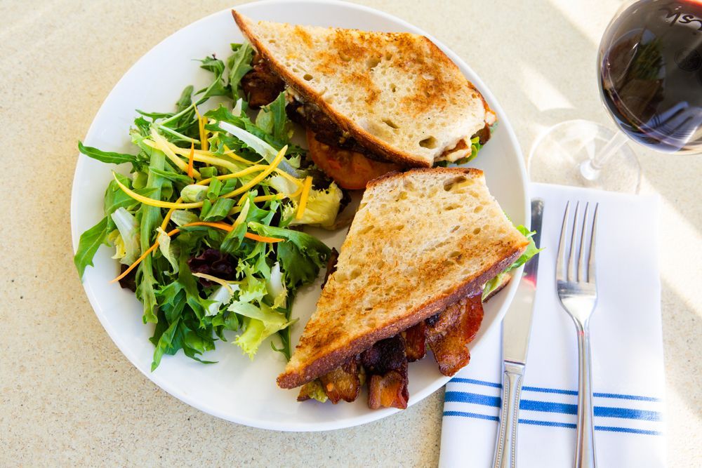 Sandwich with bacon and salad on a plate, next to a glass of red wine and silverware.
