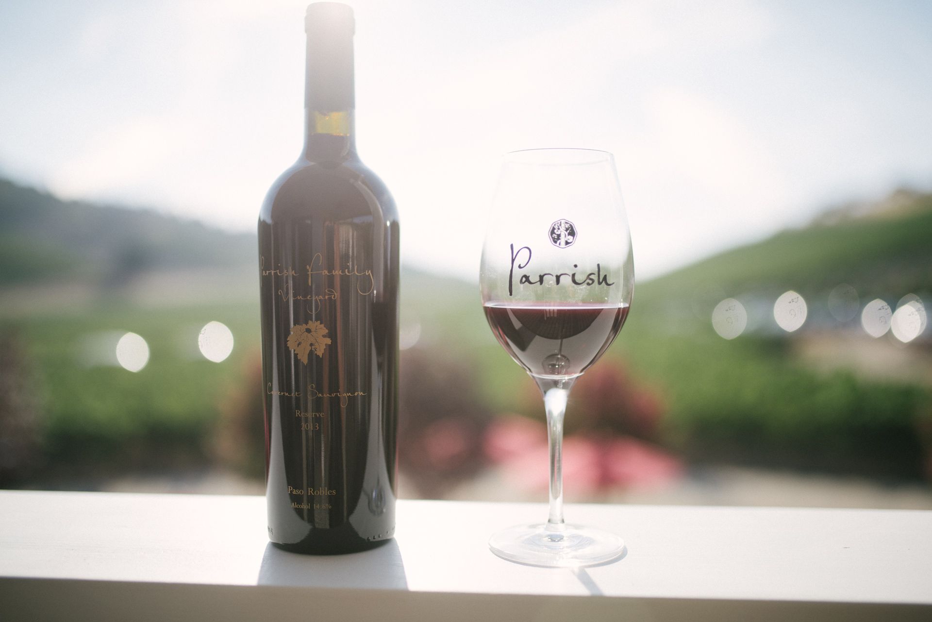 A bottle of wine and a glass of wine on a table with a blurred view of the vineyard in the back.