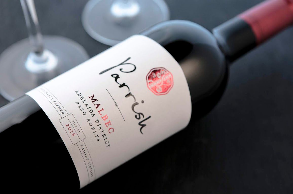 Bottle of Parrish Malbec wine with two glasses in the background. White label, dark setting.