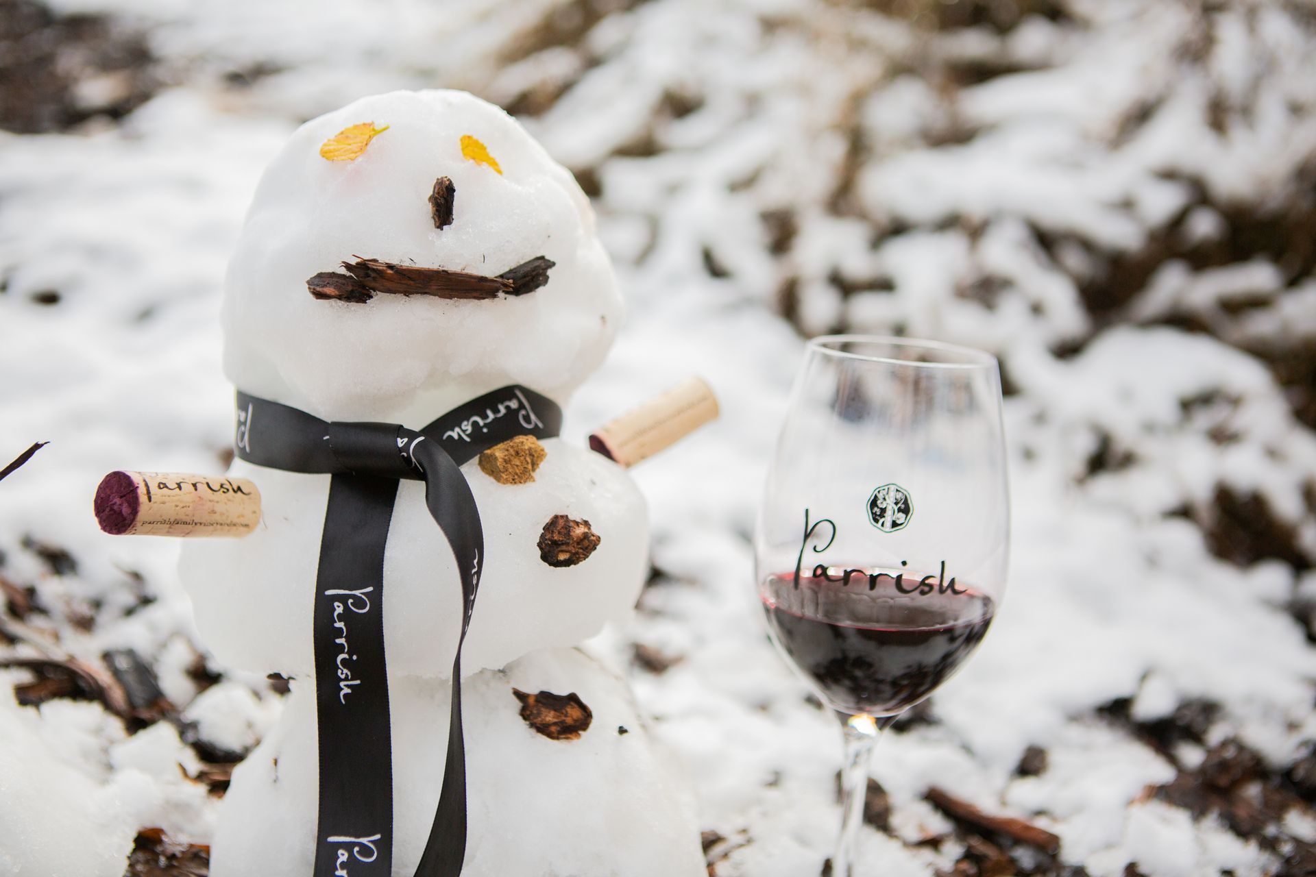 Snowman with wine cork arms next to a glass of Parrish red wine in a snowy outdoor setting.