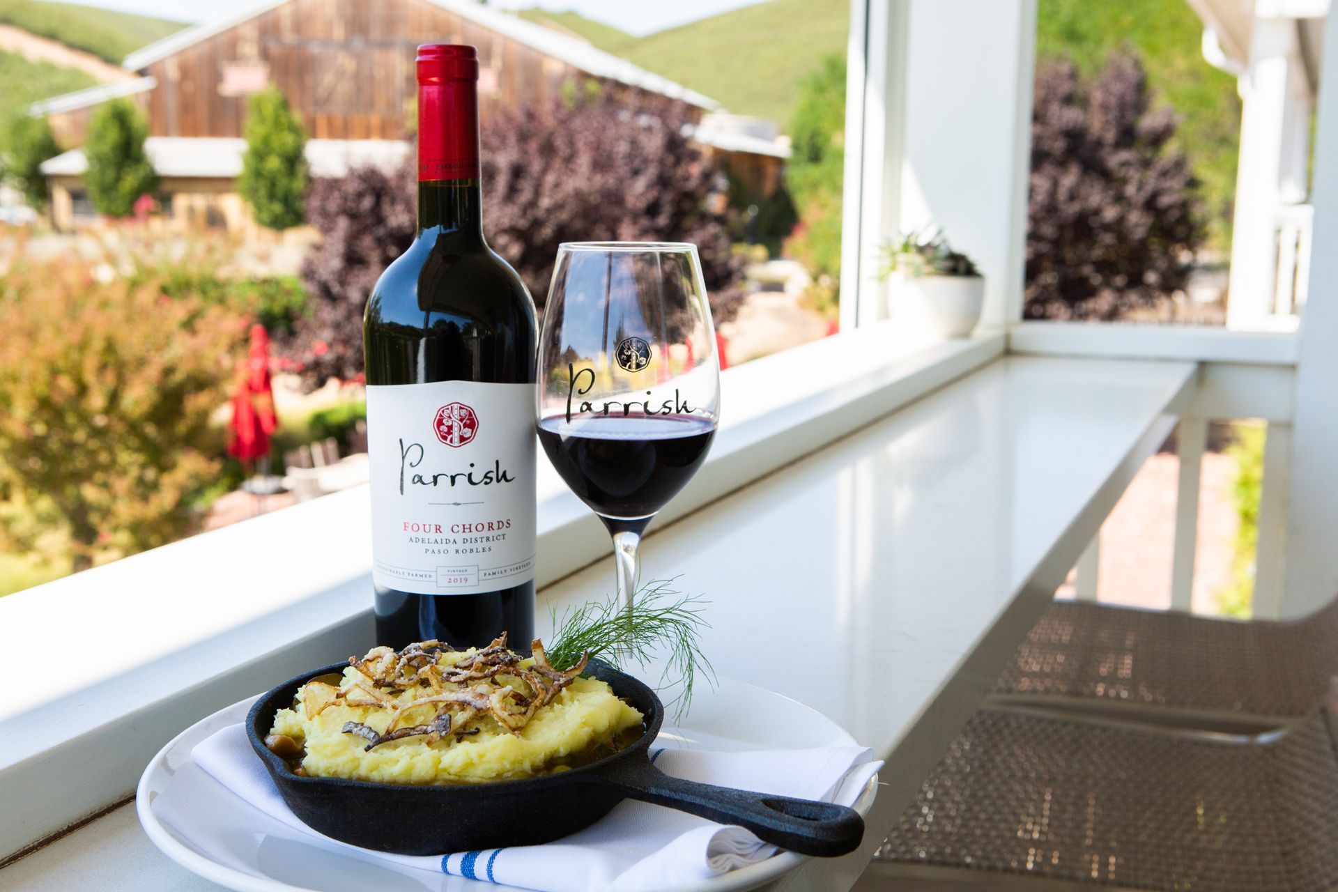 A bottle of wine and a glass of wine in front of a view of a garden.