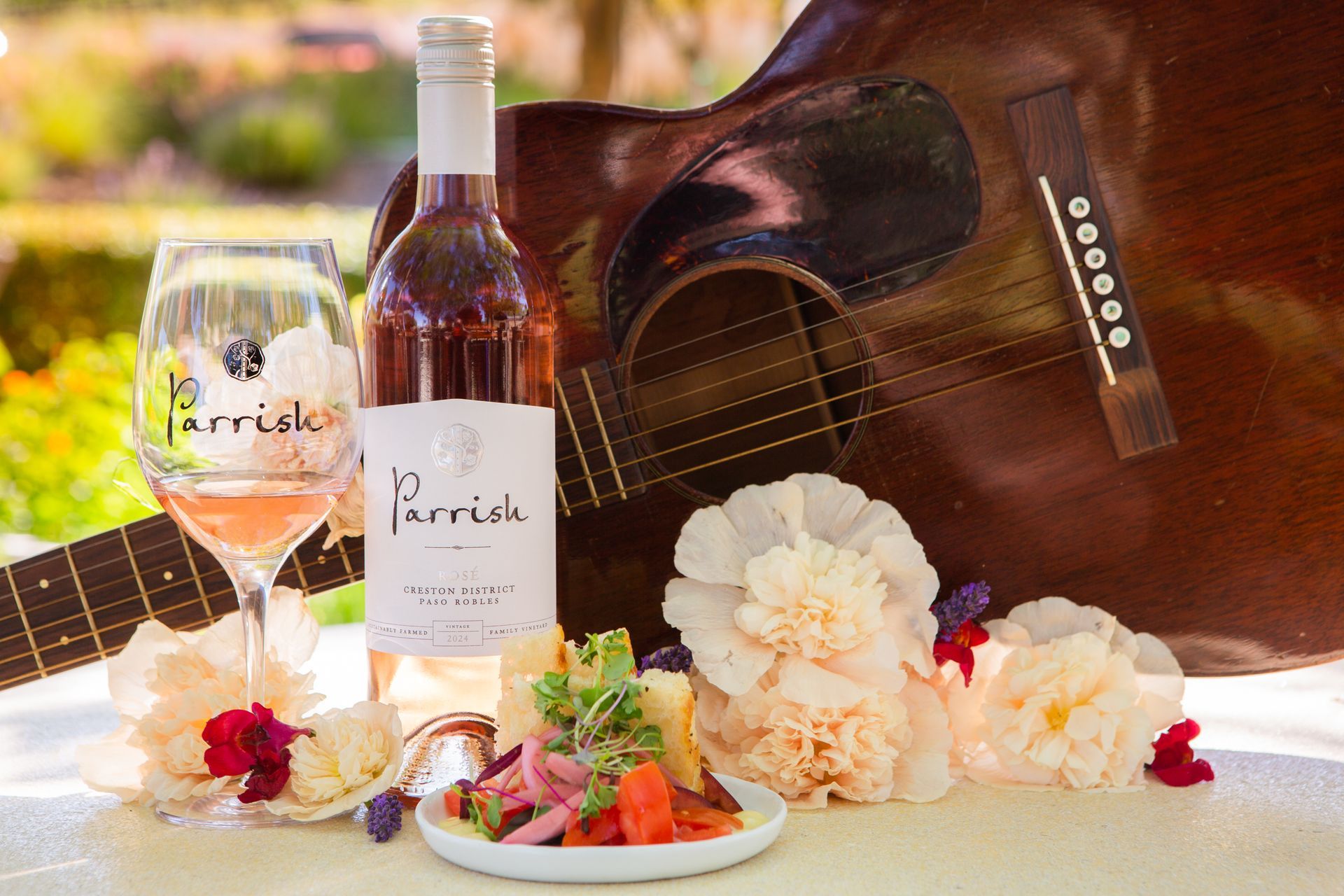 A glass of Rosé next to a bottle of Parrish Grenache Rosé, arranged aside flowers and an acoustic guitar.