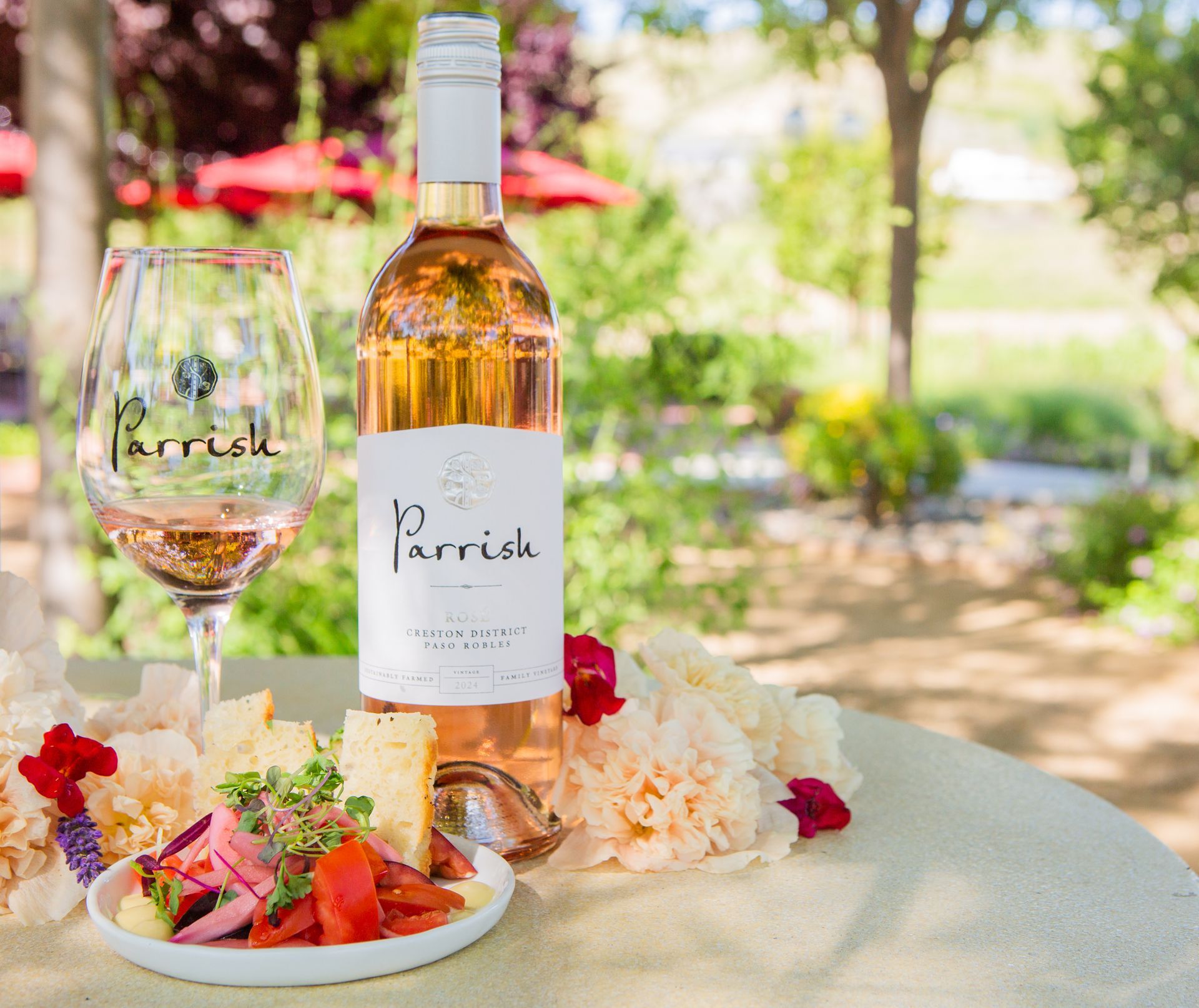 Bottle of rosé wine with glass, food, and flowers on a table outdoors.