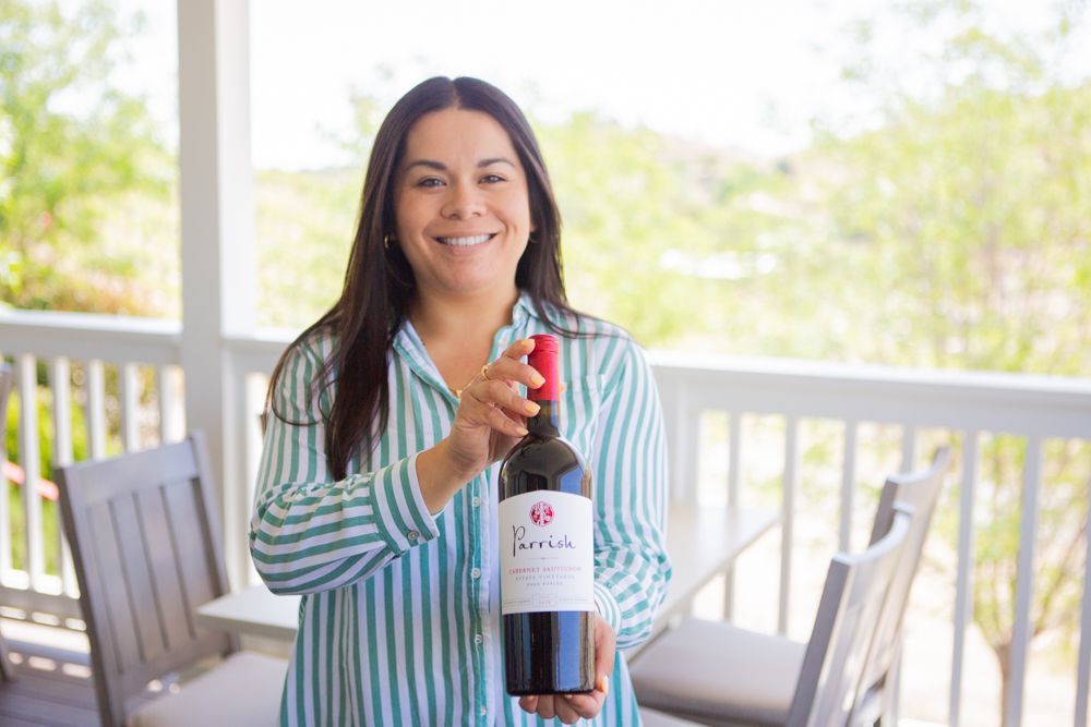 A woman is holding a bottle of Parrish red wine on a porch.