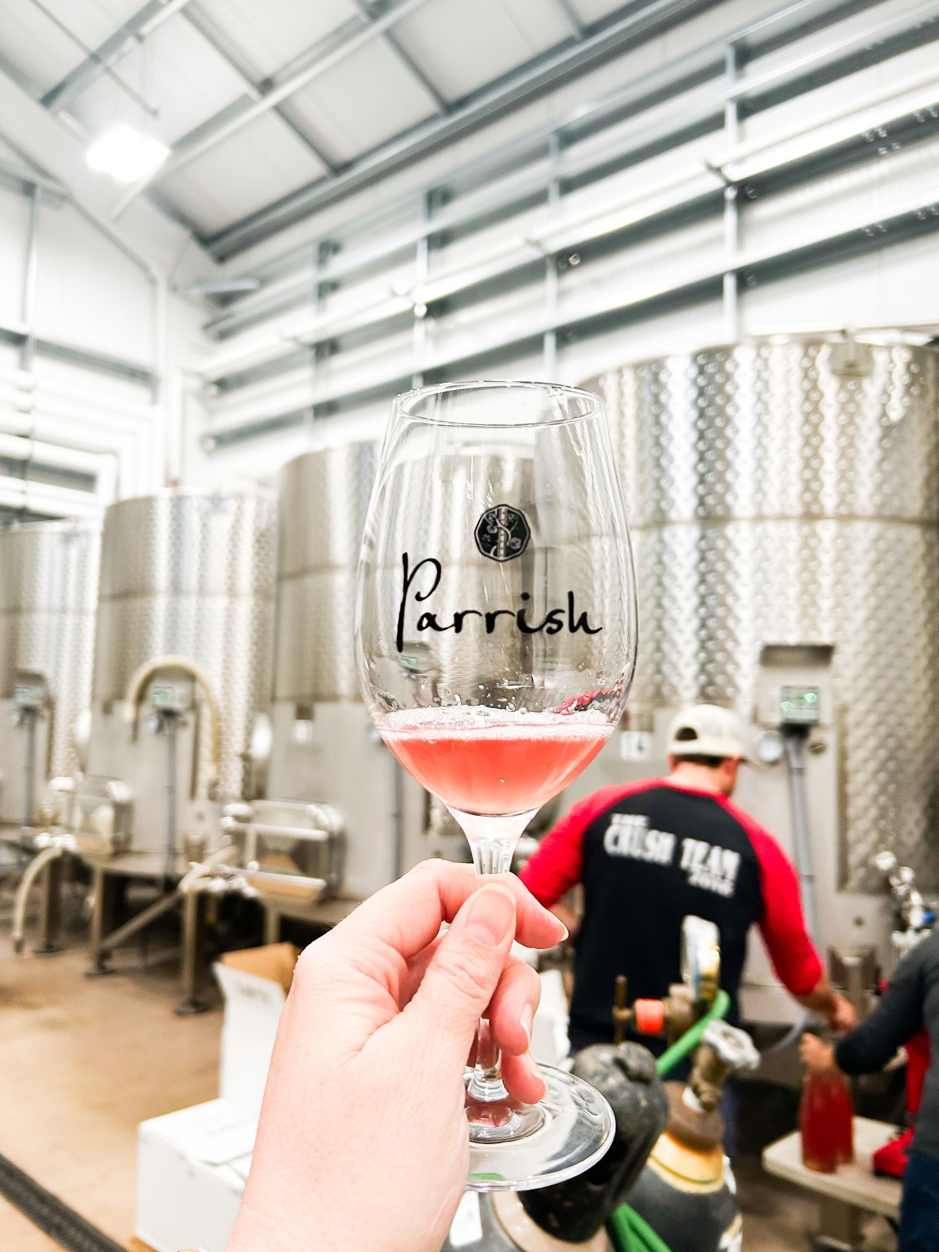 A Parrish-branded wine glass being held up in the production winery, filled with a splash of 2025 Grenache Pét-Nat. Parrish employees seen in the back working with the stainless steel tanks used to ferment the wine.