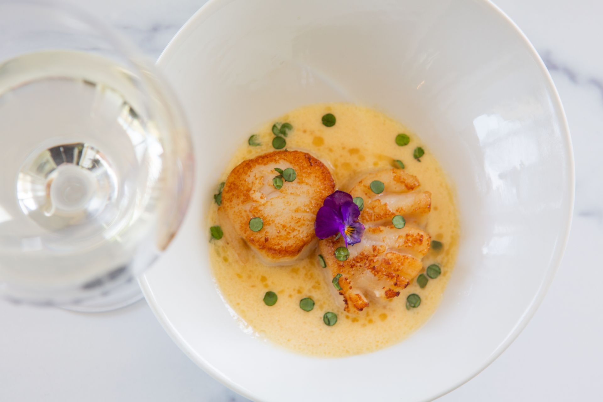 Elegant plated appetizer with creamy sauce, golden rounds, herb garnish, and a purple edible flower beside a glass of Parrish Sauvignon Blanc.