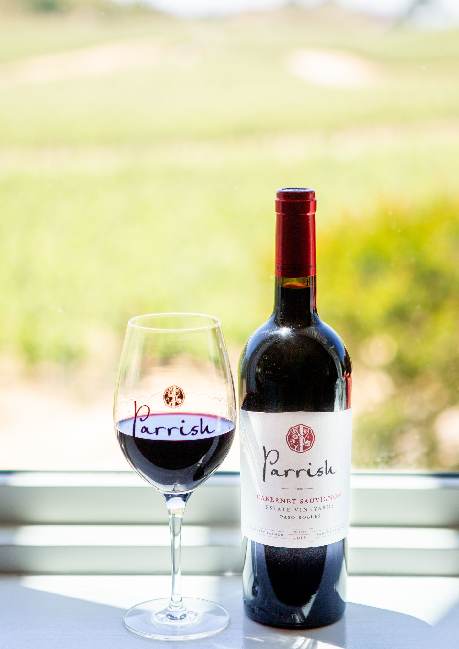 2019 Parrish Estate Cabernet Sauvignon wine bottle and glass on a balcony ledge with a blurred green vineyard beyond