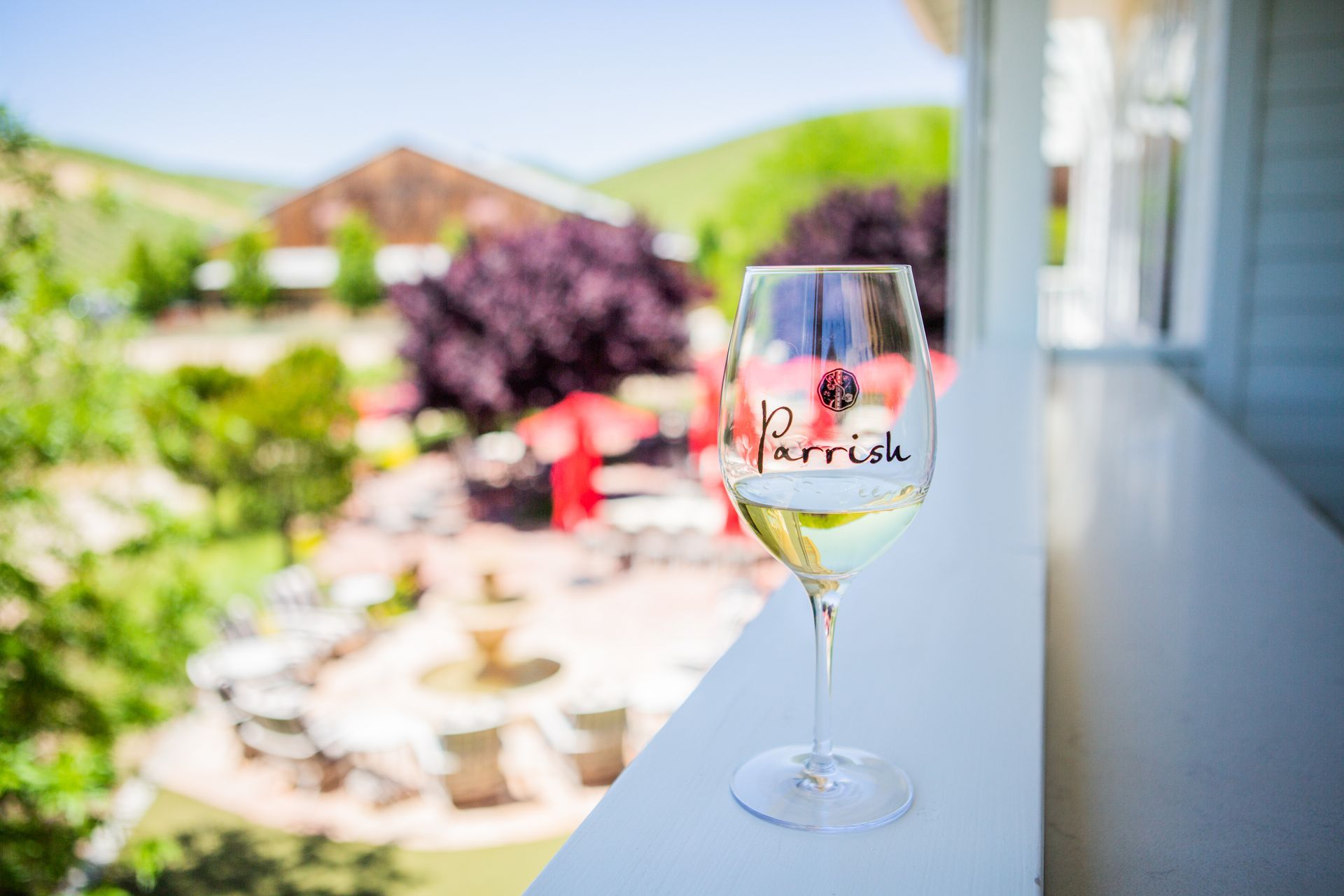 Parrish-branded wine glass on the tasting room balcony railing overlooking a sunny patio and garden, the production winery seen in the distance.