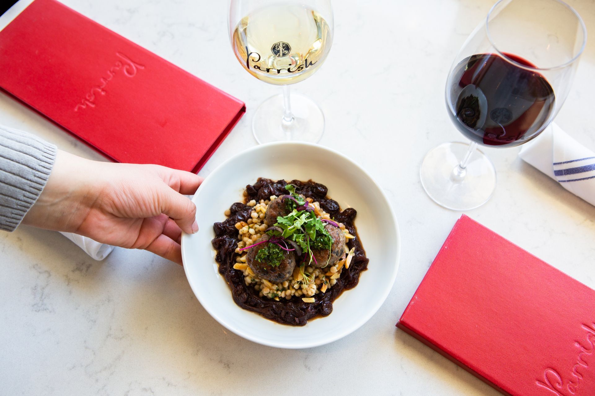 Hand reaching toward a plated dish beside red menus and two glasses of Parrish wine at a table during a wine tasting experience