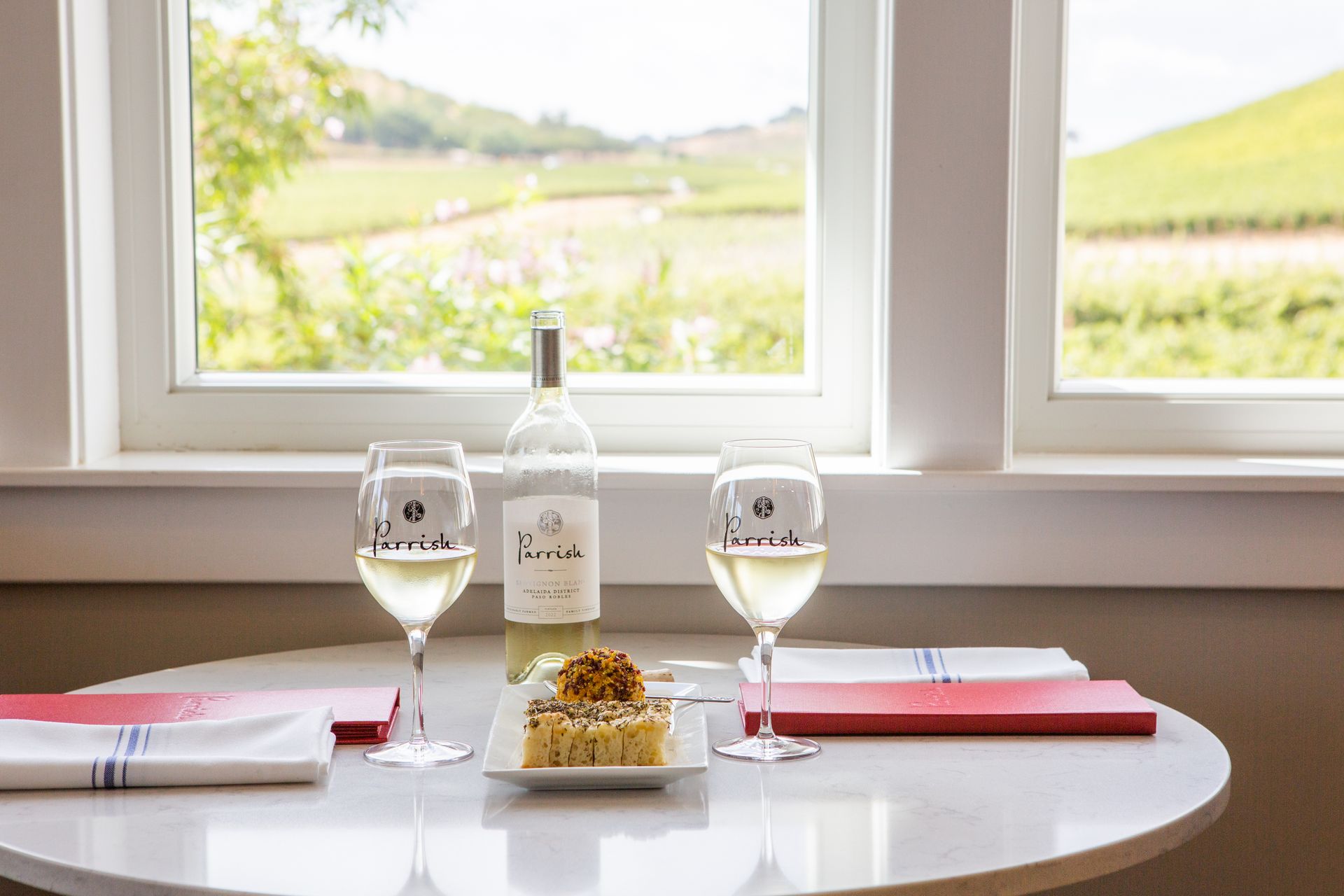 A table set for a wine tasting experience in the Parrish Family Vineyard tasting room by a window with two wine glasses and a bottle of Sauvignon Blanc wine, paired with house-made focaccia bread and butter.