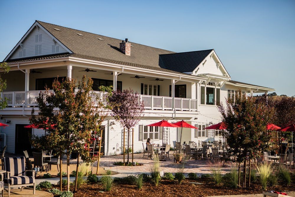 White farmhouse-style building with a two-level veranda, patio tables, red umbrellas, and trees under a blue sky.