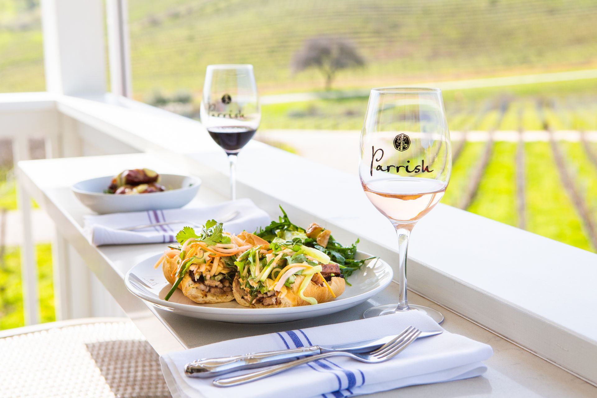 Two plated meals and wine glasses on a bright table overlooking vineyard rows