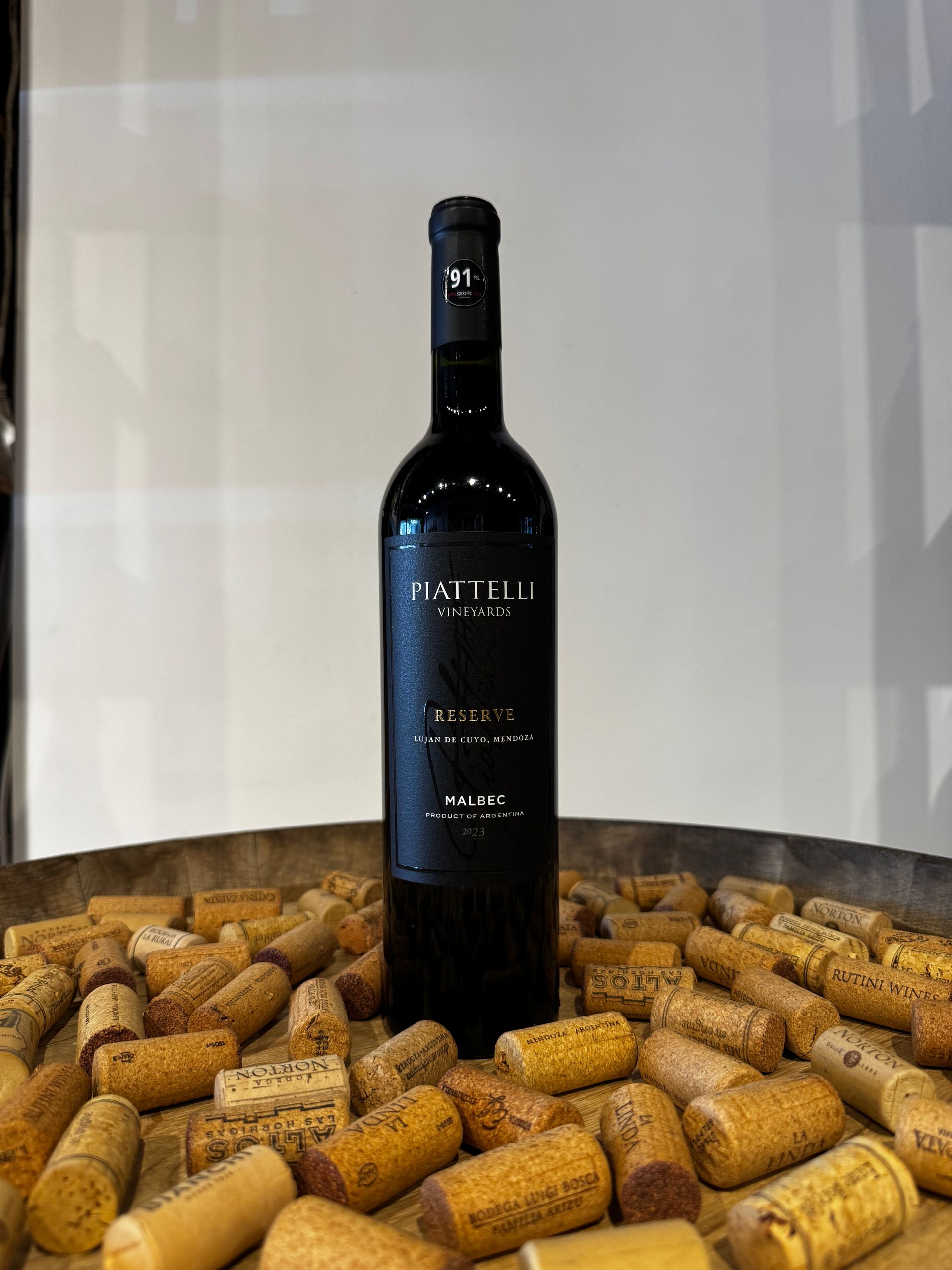 Botella de vino tinto sobre un montón de corchos, dentro de un cuenco de madera. Etiqueta negra, fondo blanco.