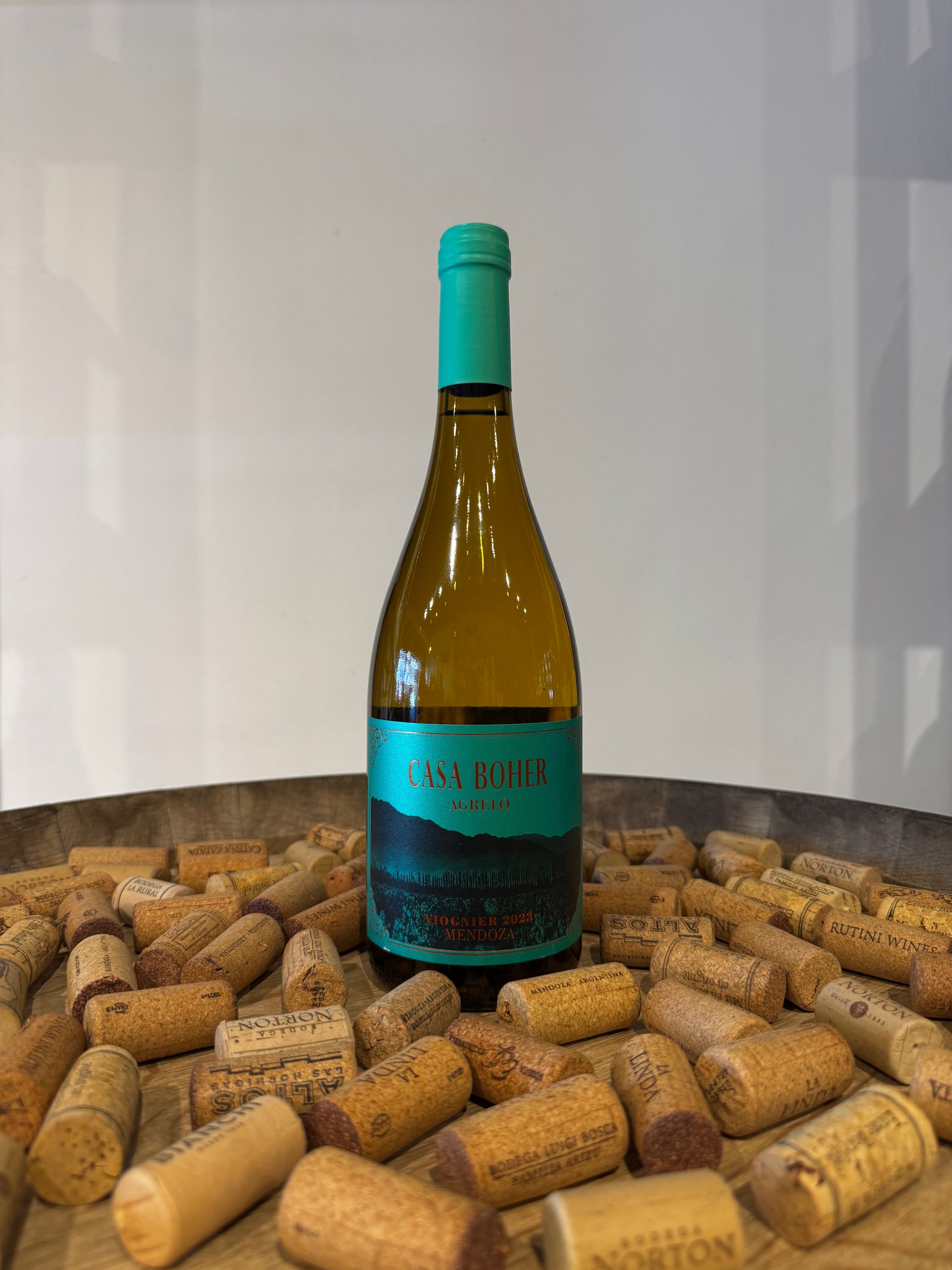 Botella de vino blanco con etiqueta verde azulado, rodeada de corchos, sobre una superficie de madera.