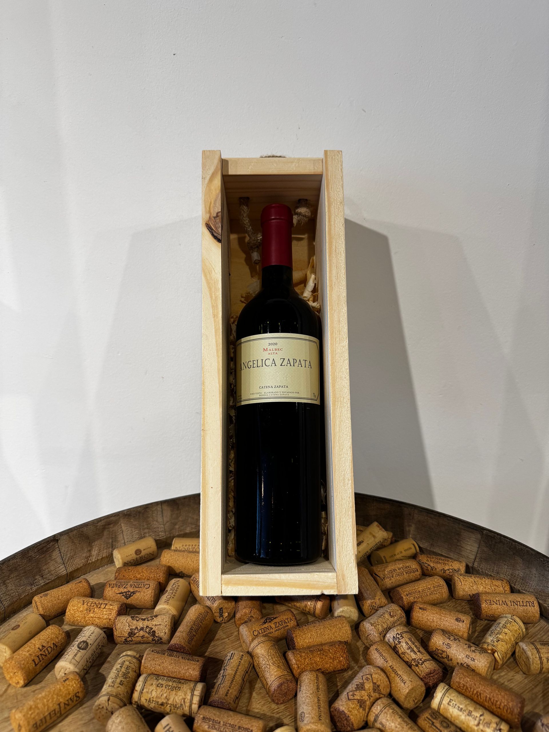 Botella de vino en una caja de madera, rodeada de corchos, sobre una superficie de madera.