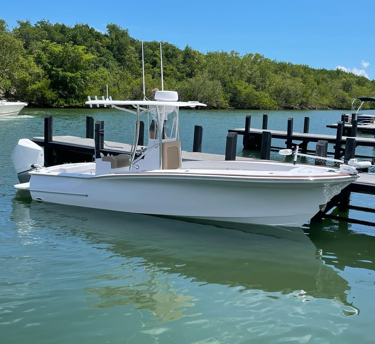 A white center-console motorboat docked at a wooden pier on calm, sunny water with lush green trees in the background.