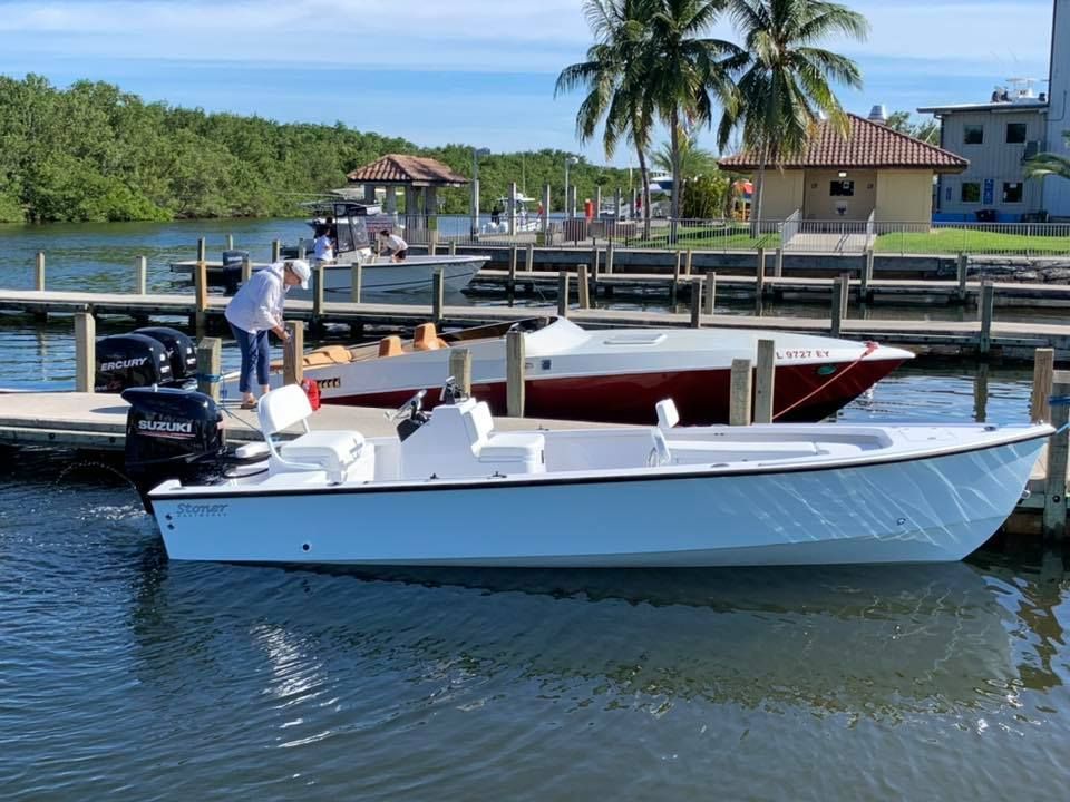 A white center-console boat docked next to a red speed boat in a marina with palm trees and a building in the background.