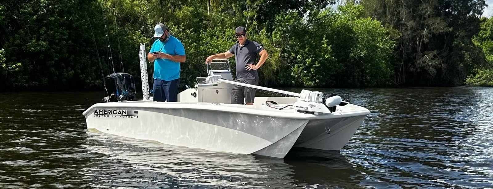 Two people stand on a white flat-bottom boat navigating a calm river lined with green trees.