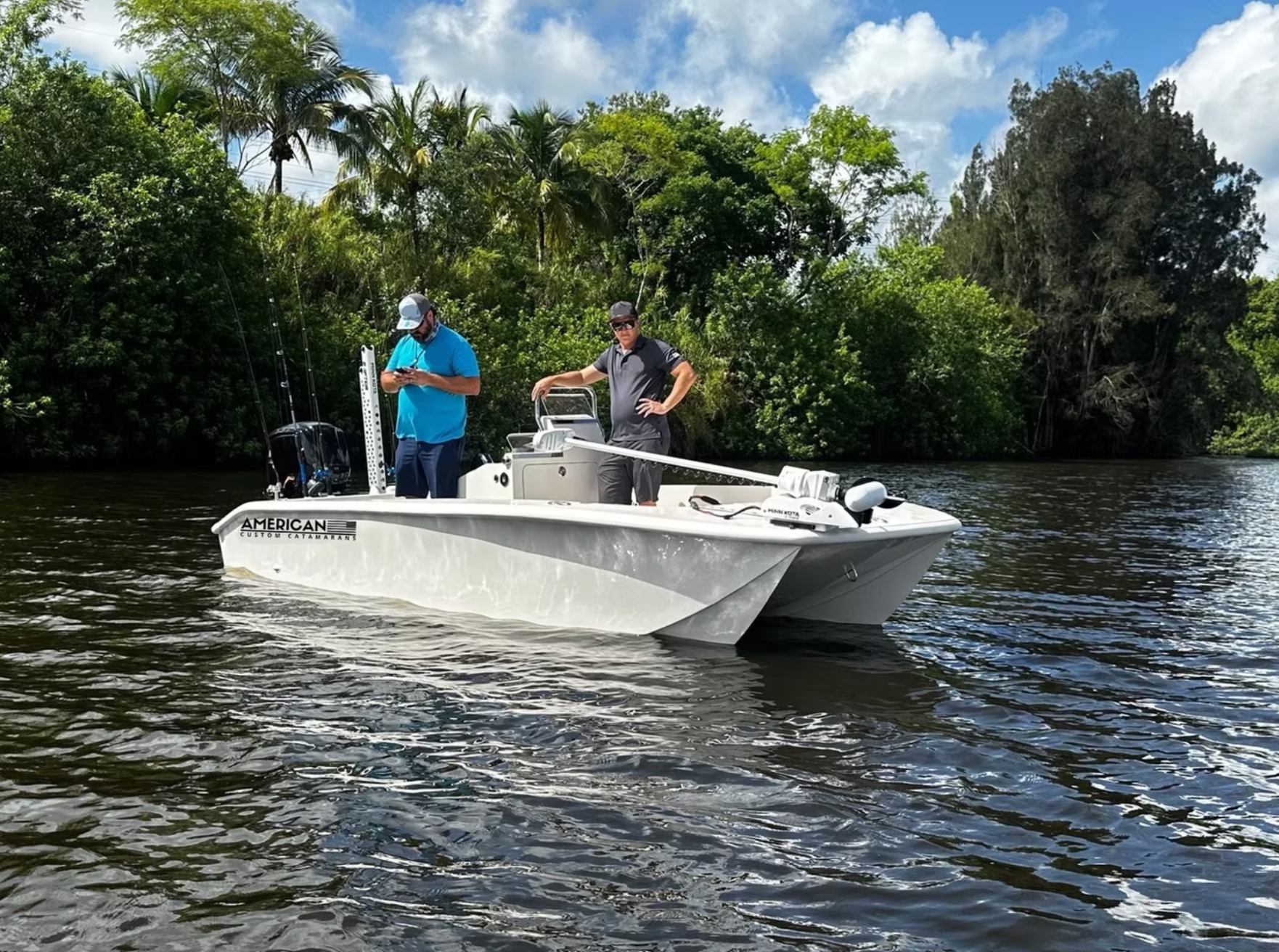 Two people on a light-colored catamaran boat in a river bordered by lush green trees under a partly cloudy sky.