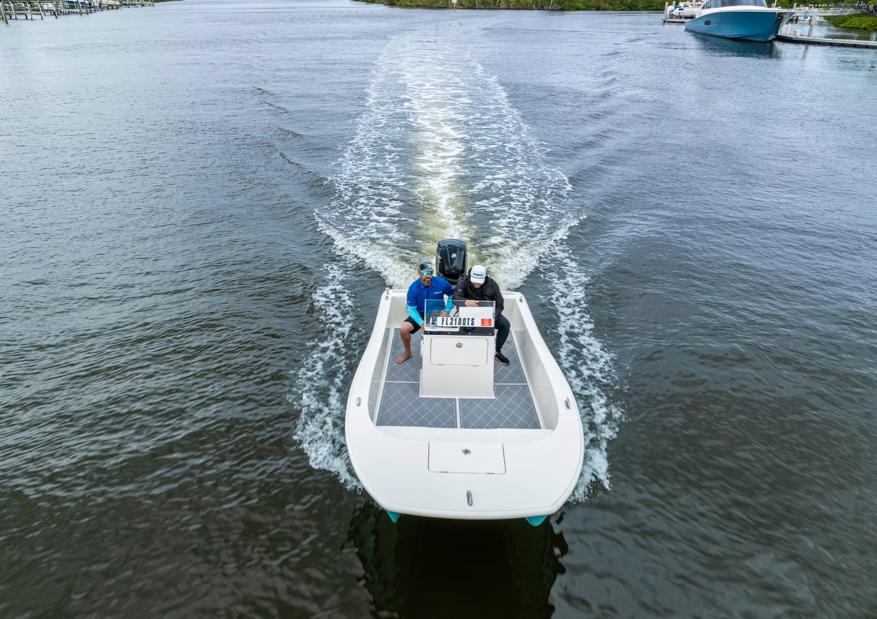 Two people steer a white center-console motorboat through calm water, leaving a bright wake behind it.