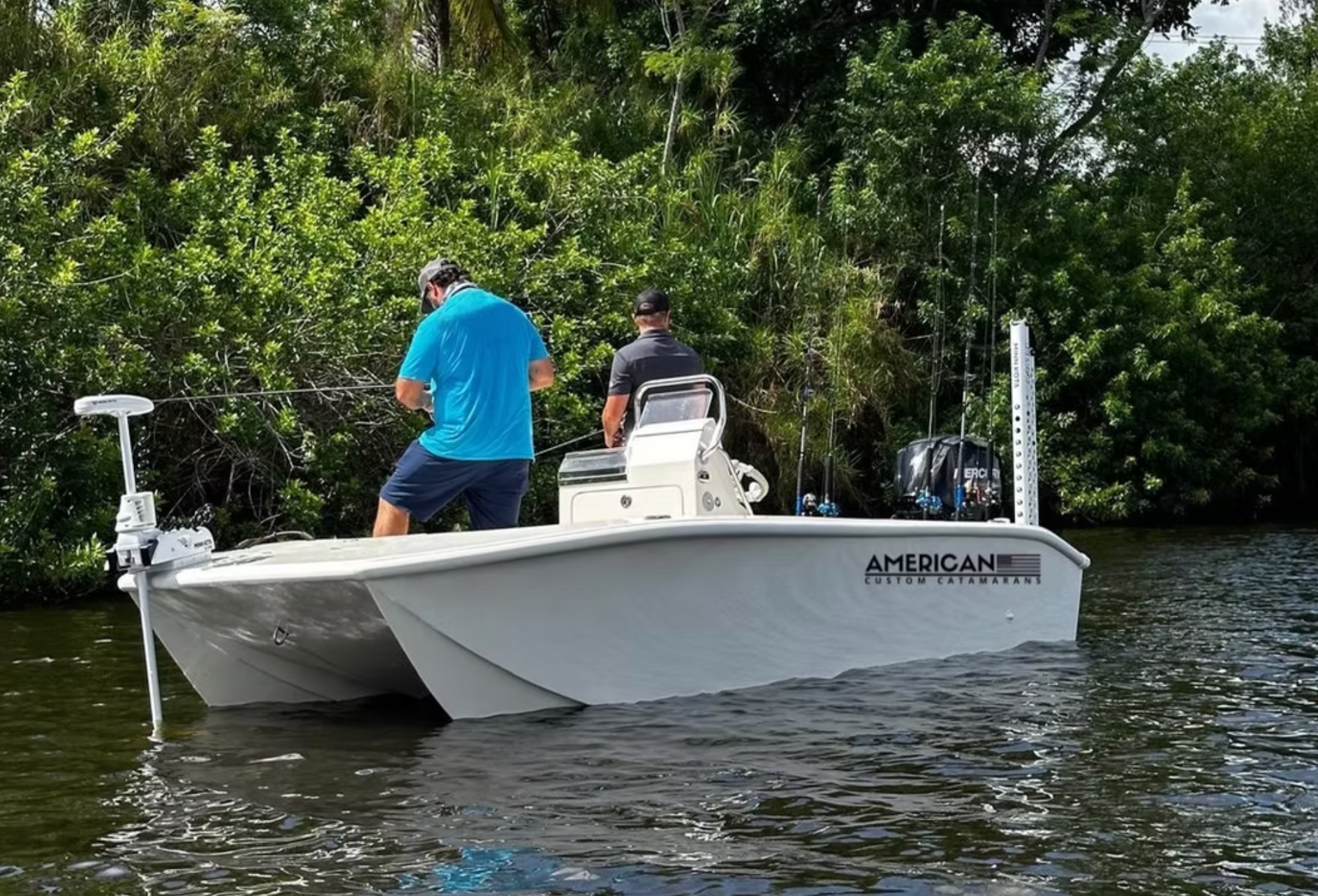 Two people fishing from a white catamaran boat in a narrow waterway lined with dense green mangroves.