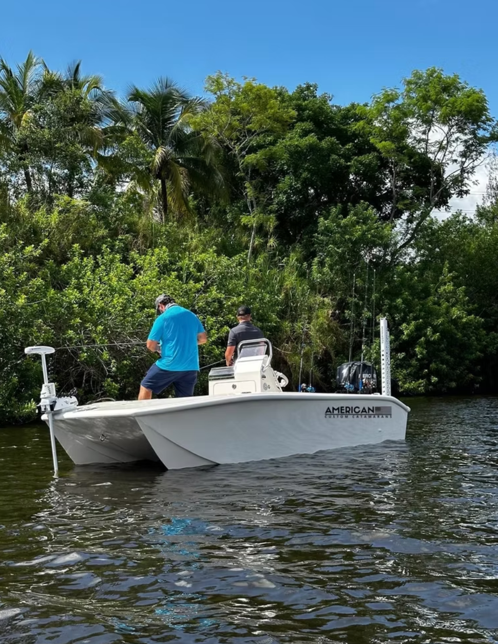 Two people fishing from a white catamaran boat in a narrow waterway surrounded by lush green trees.