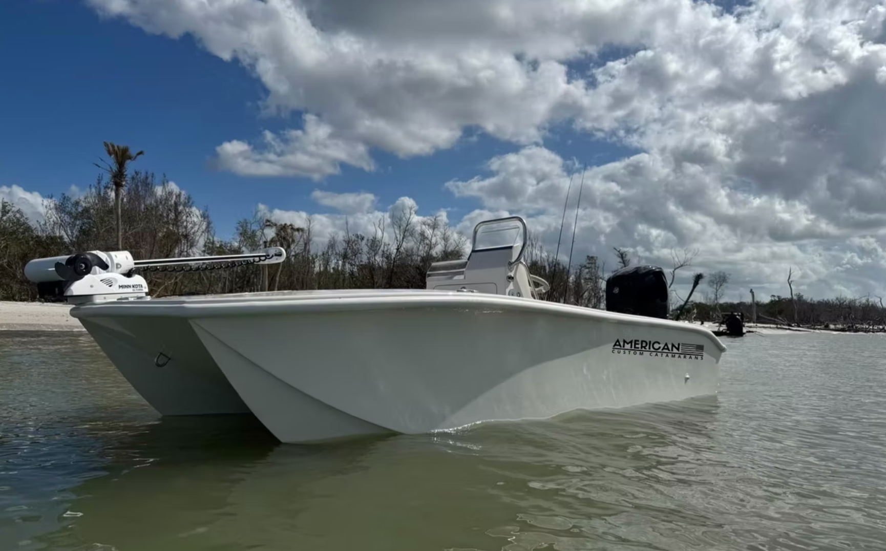 A white catamaran-style center console boat floats in shallow, calm water under a partly cloudy blue sky.