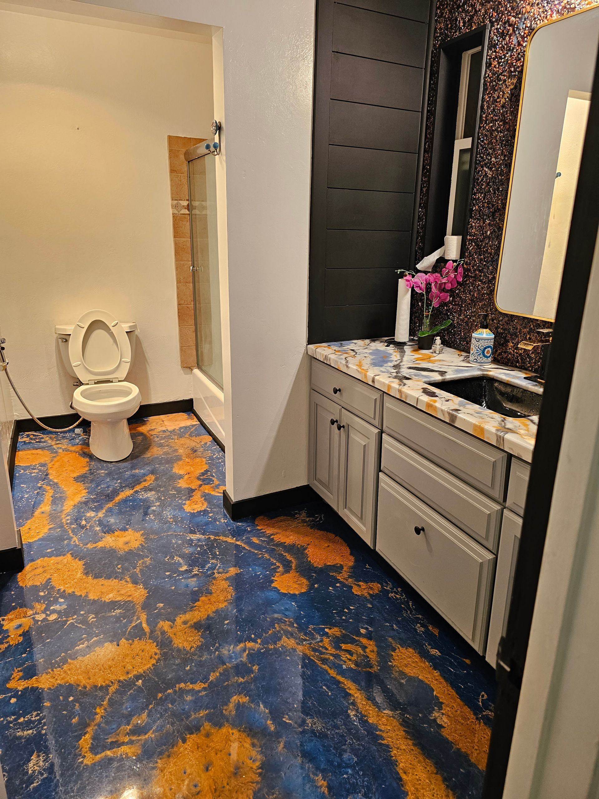 Bathroom with blue and gold epoxy floor, gray vanity, toilet, and shower. Mirror has a sparkly border.