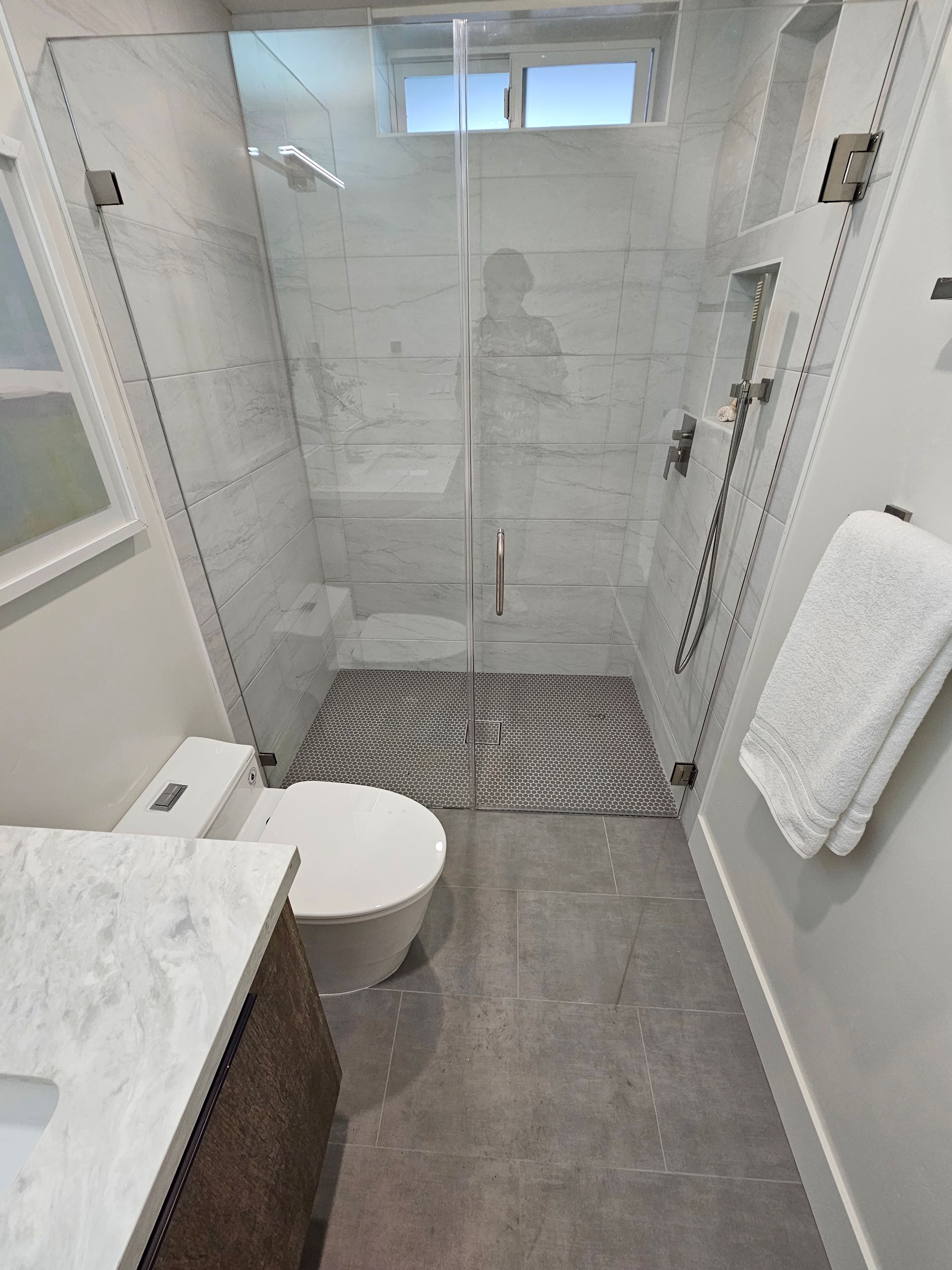 Bathroom with glass shower, white marble tile, toilet, and vanity. Gray floor and a window.