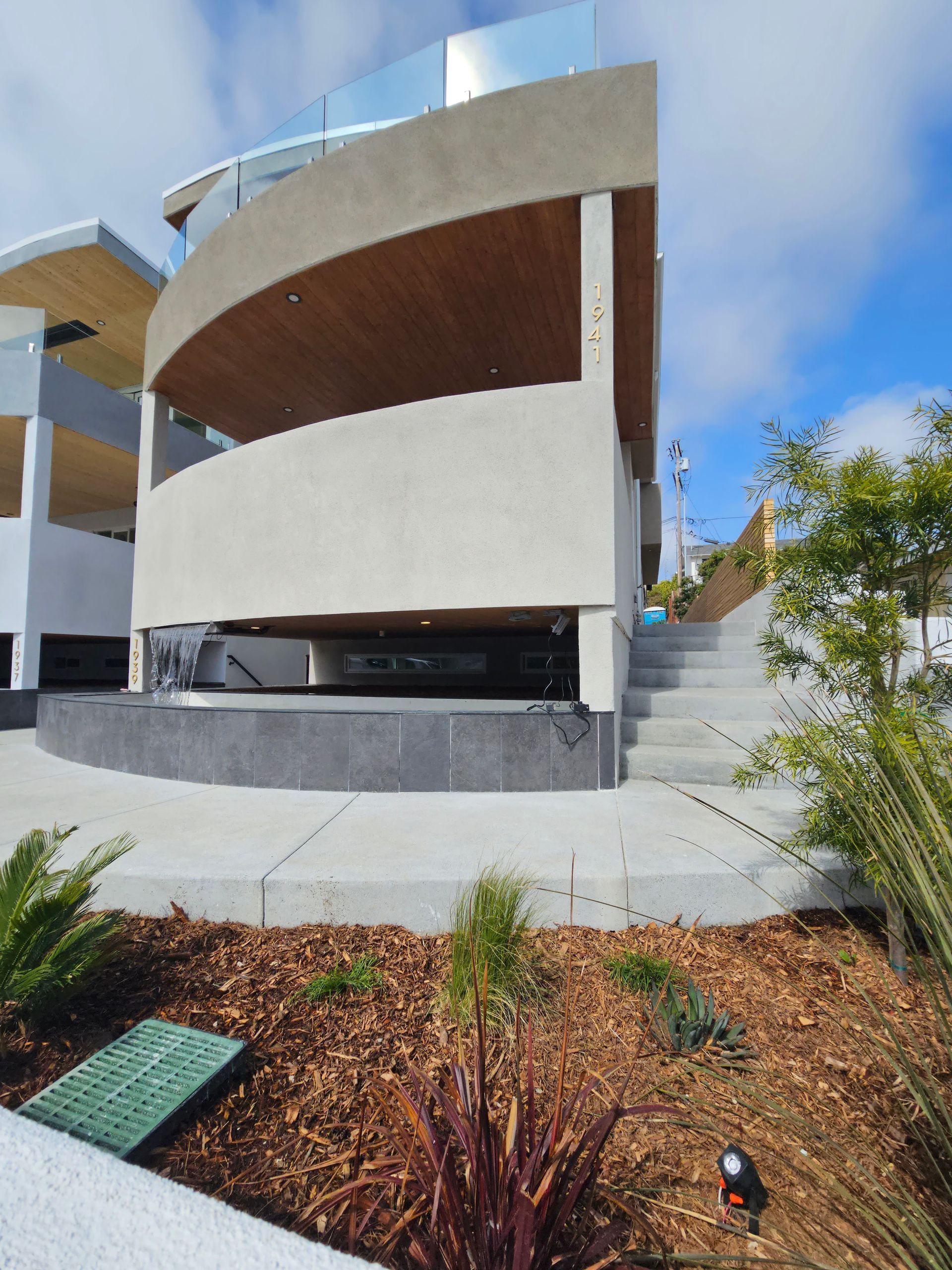 A modern building with curved, light-colored concrete exterior walls, wood-paneled balconies, and stone steps outside.