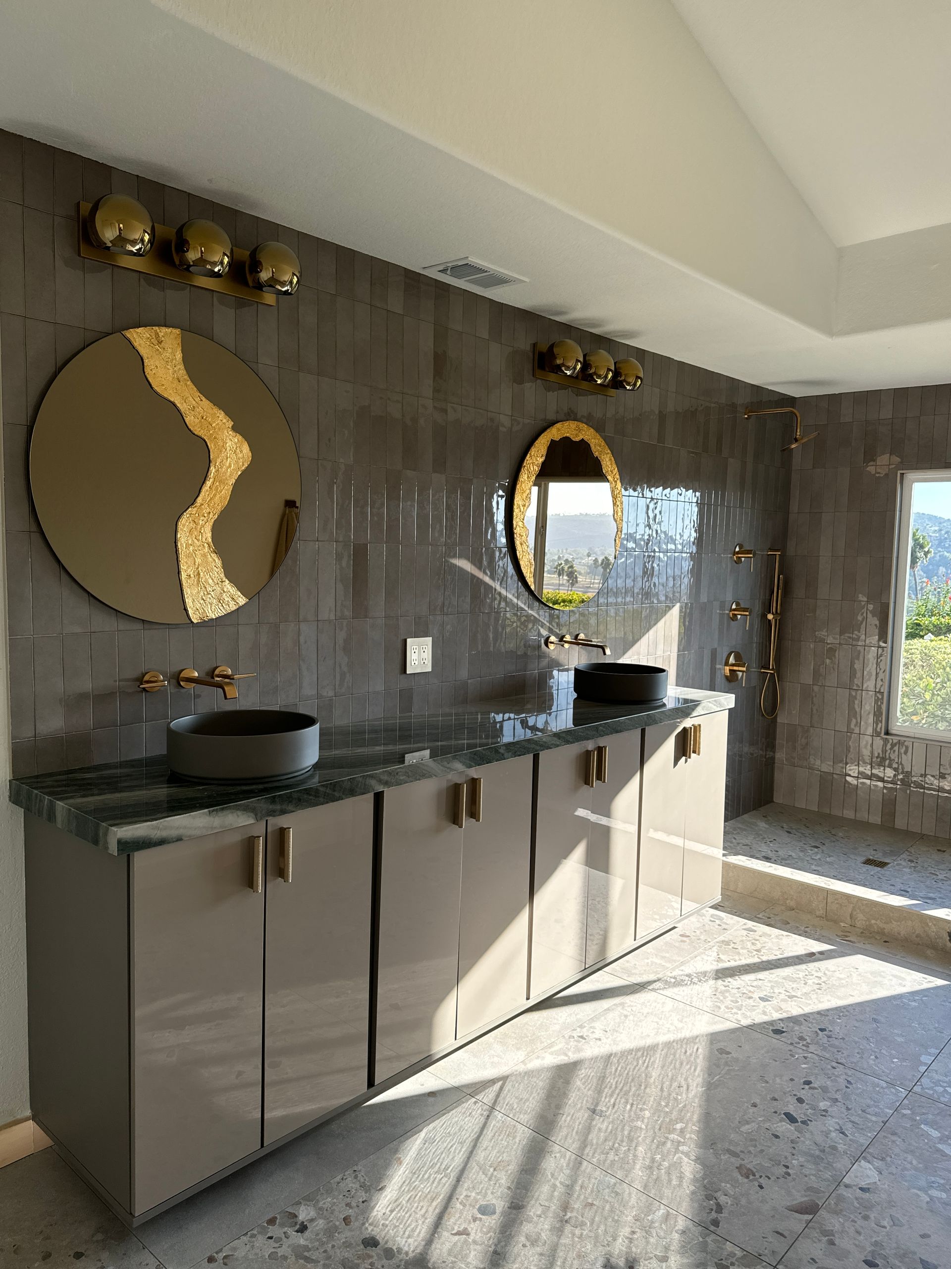 Bathroom with gray tile wall, beige cabinets, two black sinks, gold mirrors, and light fixtures.