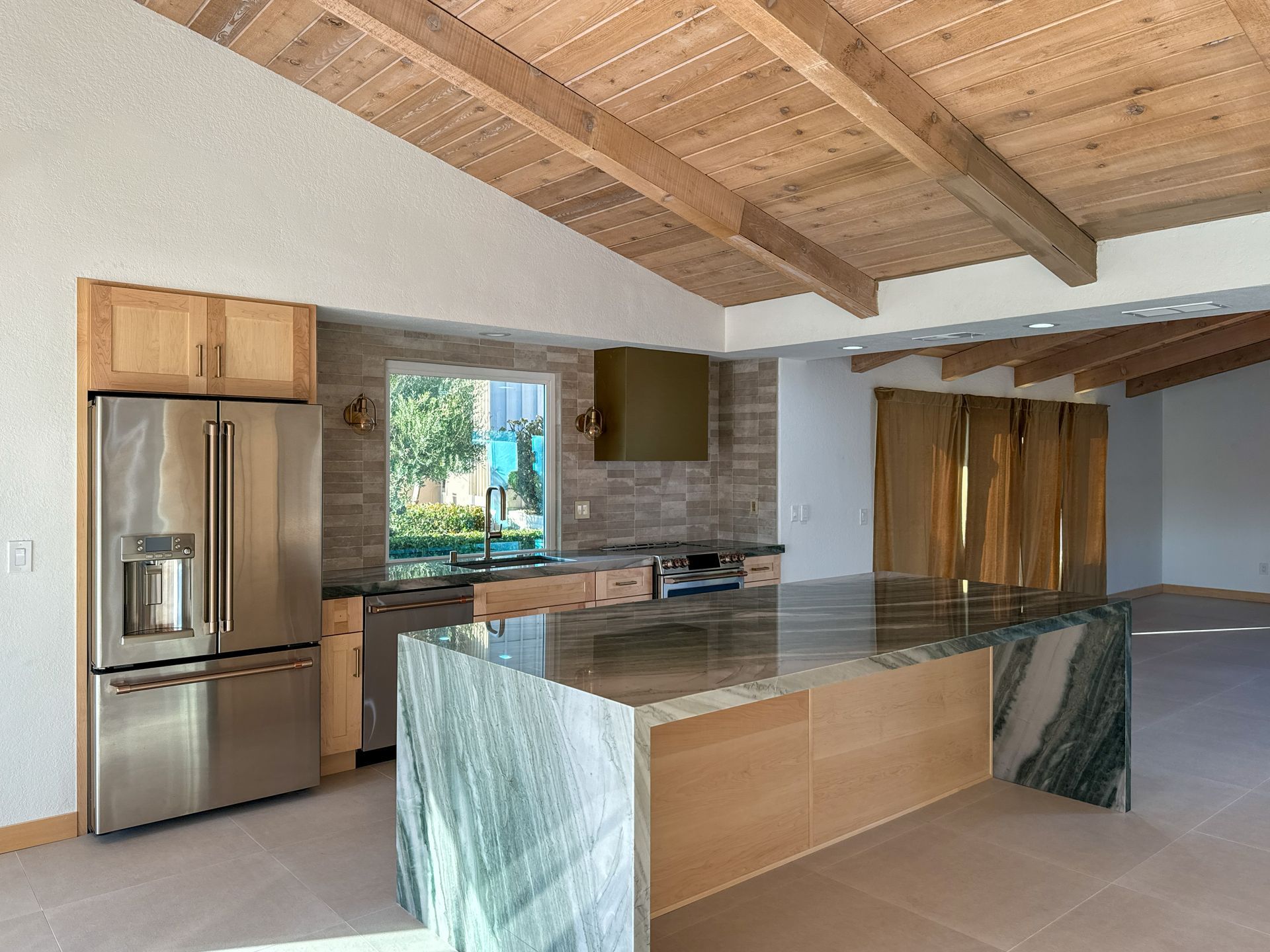 Modern kitchen with wood-beamed ceiling, quartzite island, wood cabinetry, and stainless-steel refrigerator.