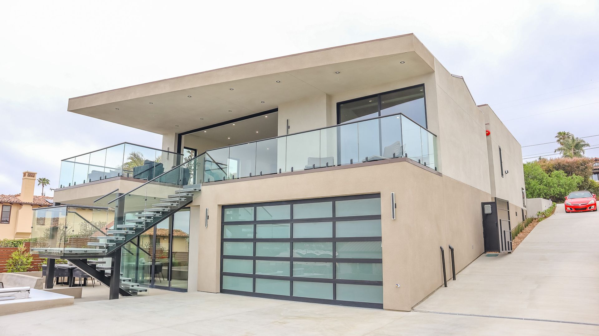 Modern two-story house with glass garage door, glass balconies, and exterior staircase on a sloped driveway.