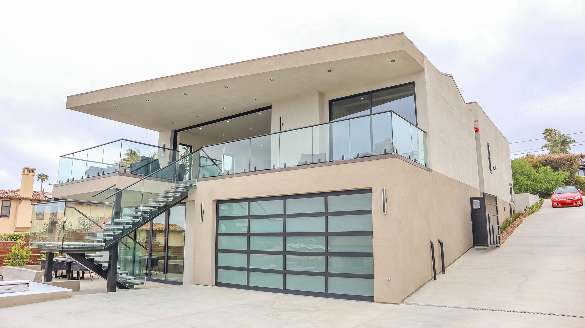 A tan, two-story modern house with a glass-railed balcony, a large frosted glass garage door, and a metal spiral staircase.