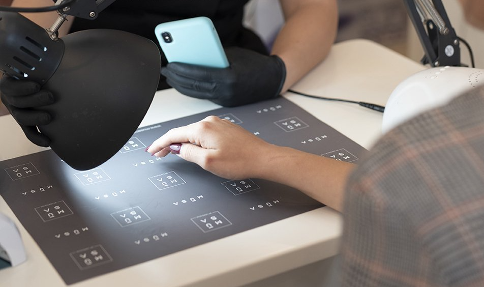 Persona che si fa fare la nail art, illuminata da una lampada, in un salone di bellezza. Un'altra persona tiene in mano un telefono.