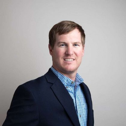 Man in a navy blazer and blue checkered shirt posing against a gray background