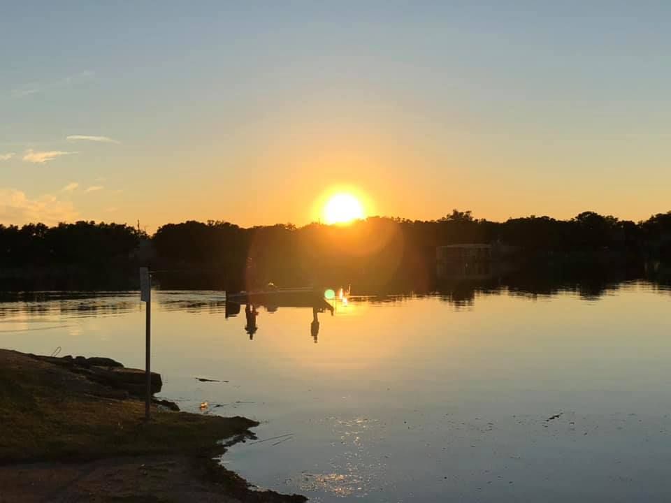 Sunset over calm water reflecting the golden sun and silhouetted trees.